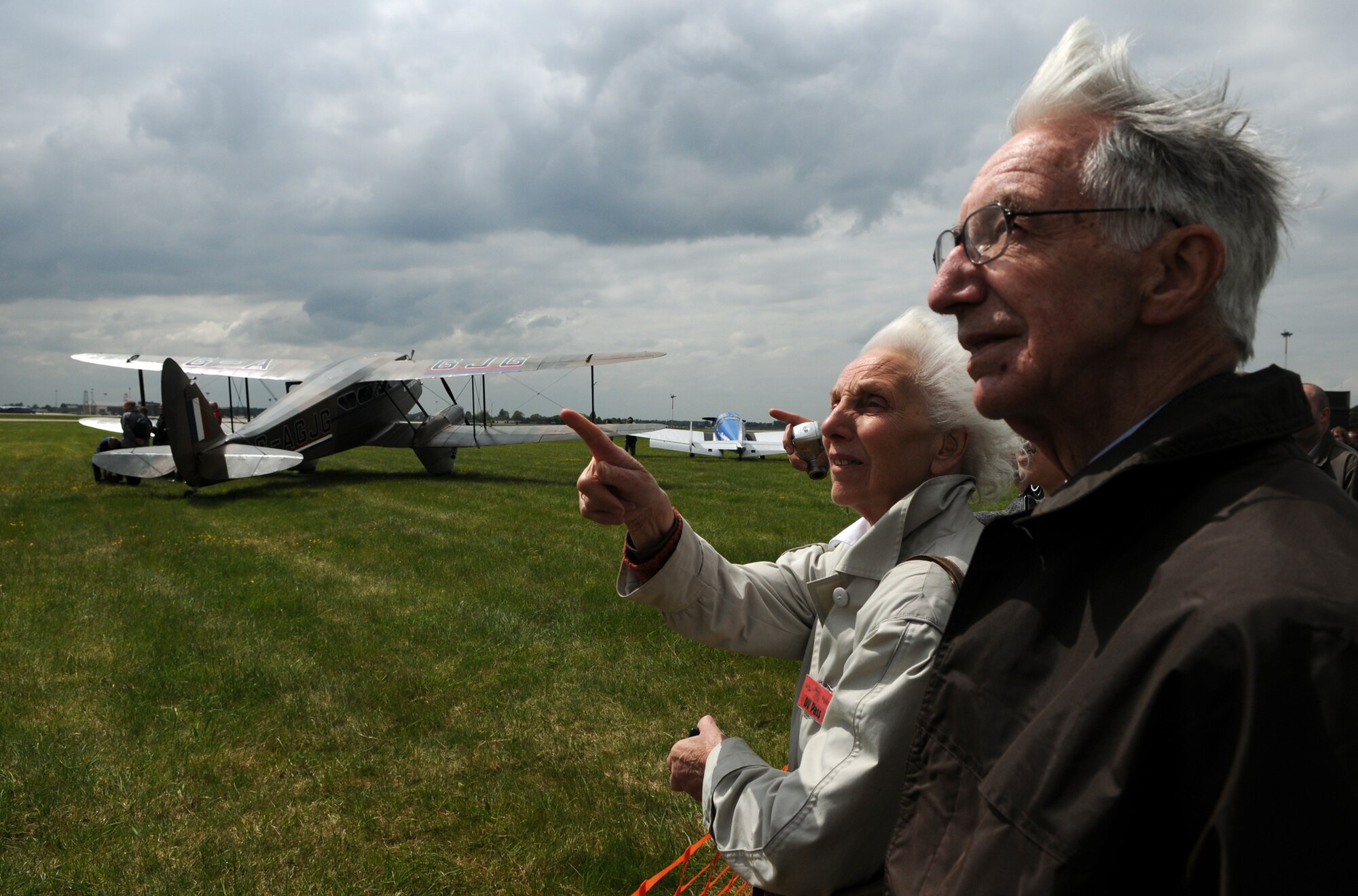 Former Royal Air Force Airman 1st Class Charlie Woolford, now 88, stands at the edge of the RAF Mildenhall flight line with Natalie Ebbs, whose father and grandfather helped build the original hangars here.  Ms. Ebbs points at one of about 20 ’30s-era aircraft which later departed the base in a recreation of the historical 1934 England to Australia Air Race during the base’s 75th Anniversary Celebration May 15.  (U.S. Air Force photo by Staff Sgt. Austin M. May)