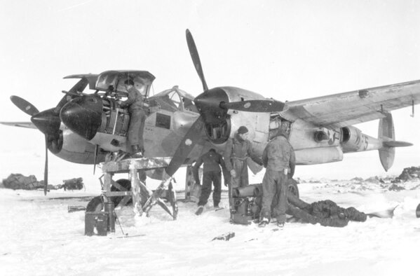 Rough field operations caused difficult working conditions for 9th Air Force maintenance personnel. Here a crew prepares to use an external heater to warm a P-38’s frozen engine so it can be started while an armorer works on the guns in the nose while atop a makeshift stand. (U.S. Air Force photo)