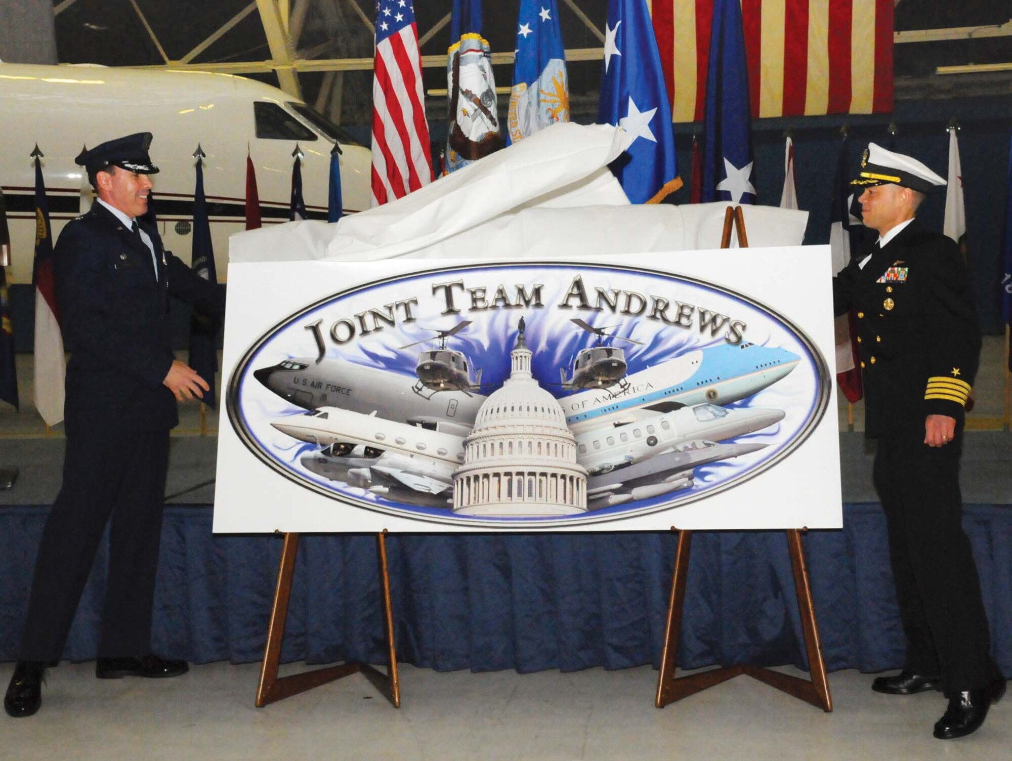 Colonel Steven M. Shepro, 316th Wing/Joint Base Andrews commander, left, and Navy Capt. Timothy Fox, Naval Air Facility Washington commanding officer, right, unveil the Joint Team Andrews logo during a Joint Base ceremony held at Hangar 3 Thursday. The event signified the unification of Andrews Air Force Base and Naval Air Facility Washington, and renamed the installation as Joint Base Andrews Naval Air Facility Washington. Among the official guests in attendance were Maj. Gen. Ralph J. Jodice II, Air Force District of Washington commander, Rear. Adm. Julius S. Caesar, Navy Installation Command Reserve deputy commander, and Navy Capt. Catherine Hanft, Naval Support Activity South Potomac commanding officer. The logo, designed by Airman 1st Class Kat Lynn Justen, 316 WG public affairs specialist, signifies the diverse missions of Joint Team Andrews and the role of the installation in the National Capital Region. (U.S. Air Force photo/ Bobby Jones)