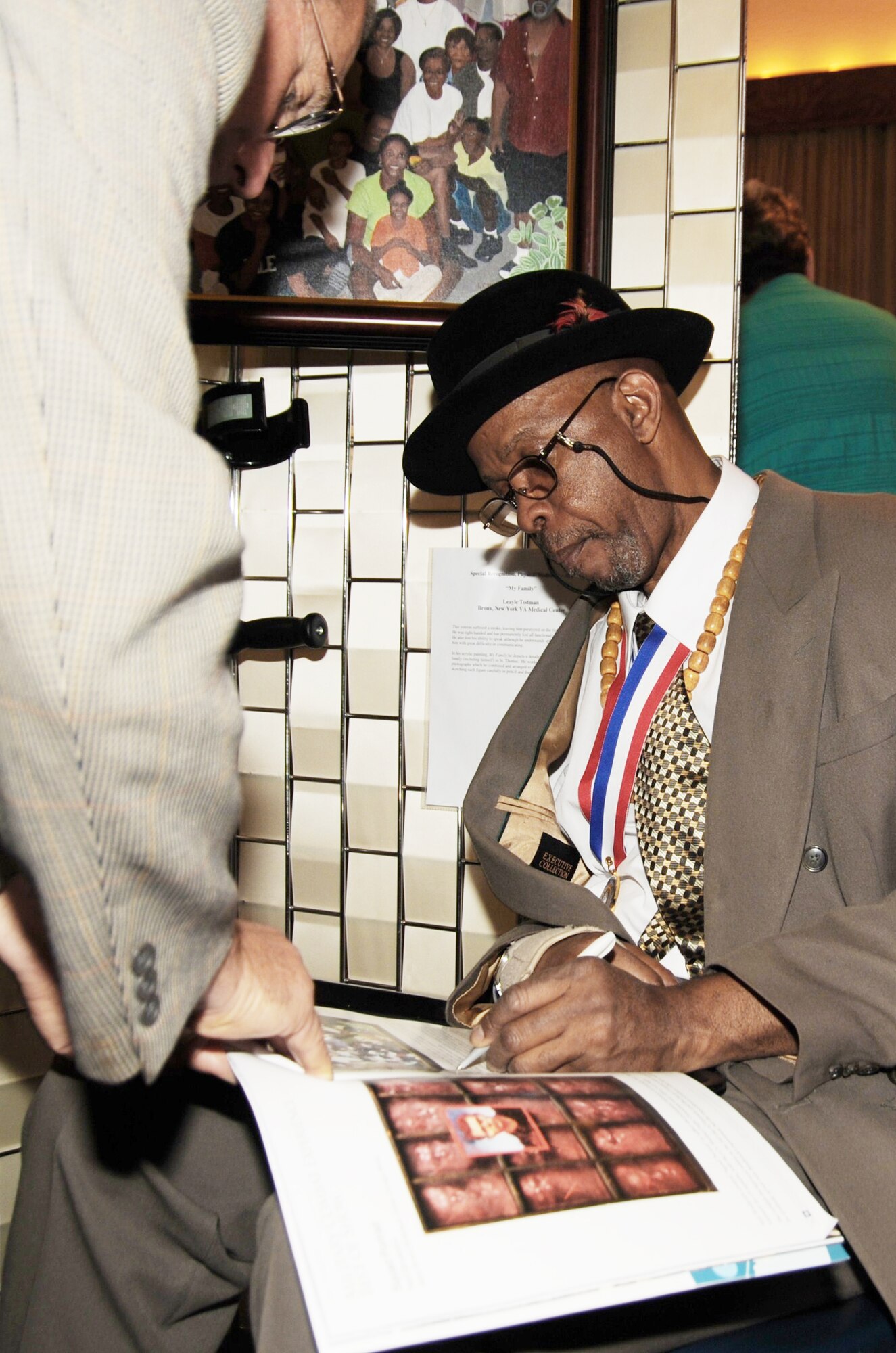 Army veteran Leayle Todman signs a photograph of his artwork for an individual  during the 2009 National Veterans Creative Arts Festival Oct. 11, 2009, at the San Antonio Municipal Auditorium in San Antonio. Mr. Todman suffered a stroke, but was still able to paint a detailed portrait of his family displayed behind him. 