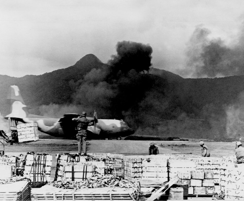 A cargo aircraft sits on a runway in front of a plume of black smoke as a man in the foreground stands on a set of pallets.