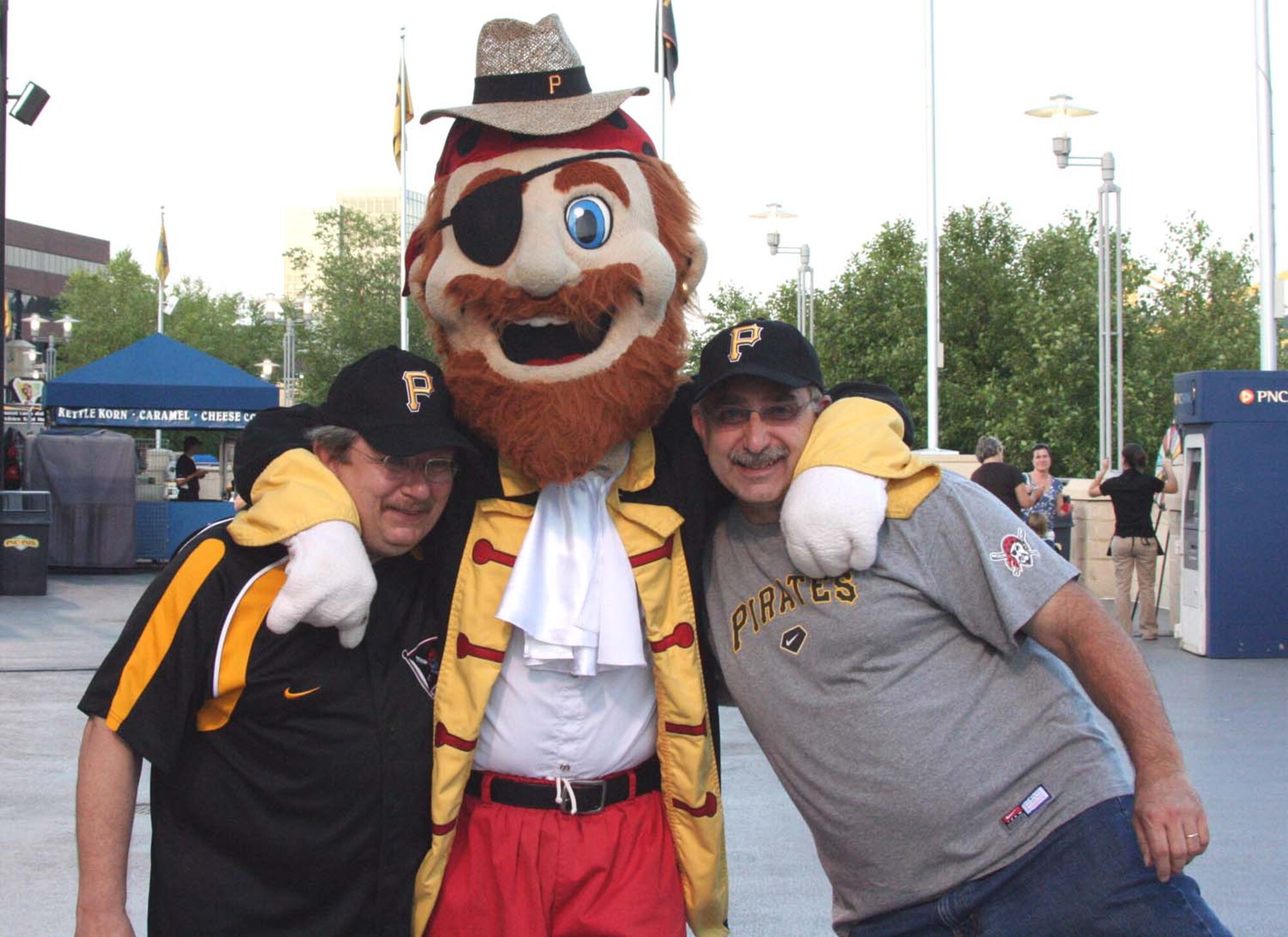 Civilian employees, Hugh Montgomery (left) and Ron Piche (right) are pulled in for a photo by their new friend, unit members on the field at PNC Park during a pregame mass enlistment ceremony Pirates mascot, Jolly Roger. 