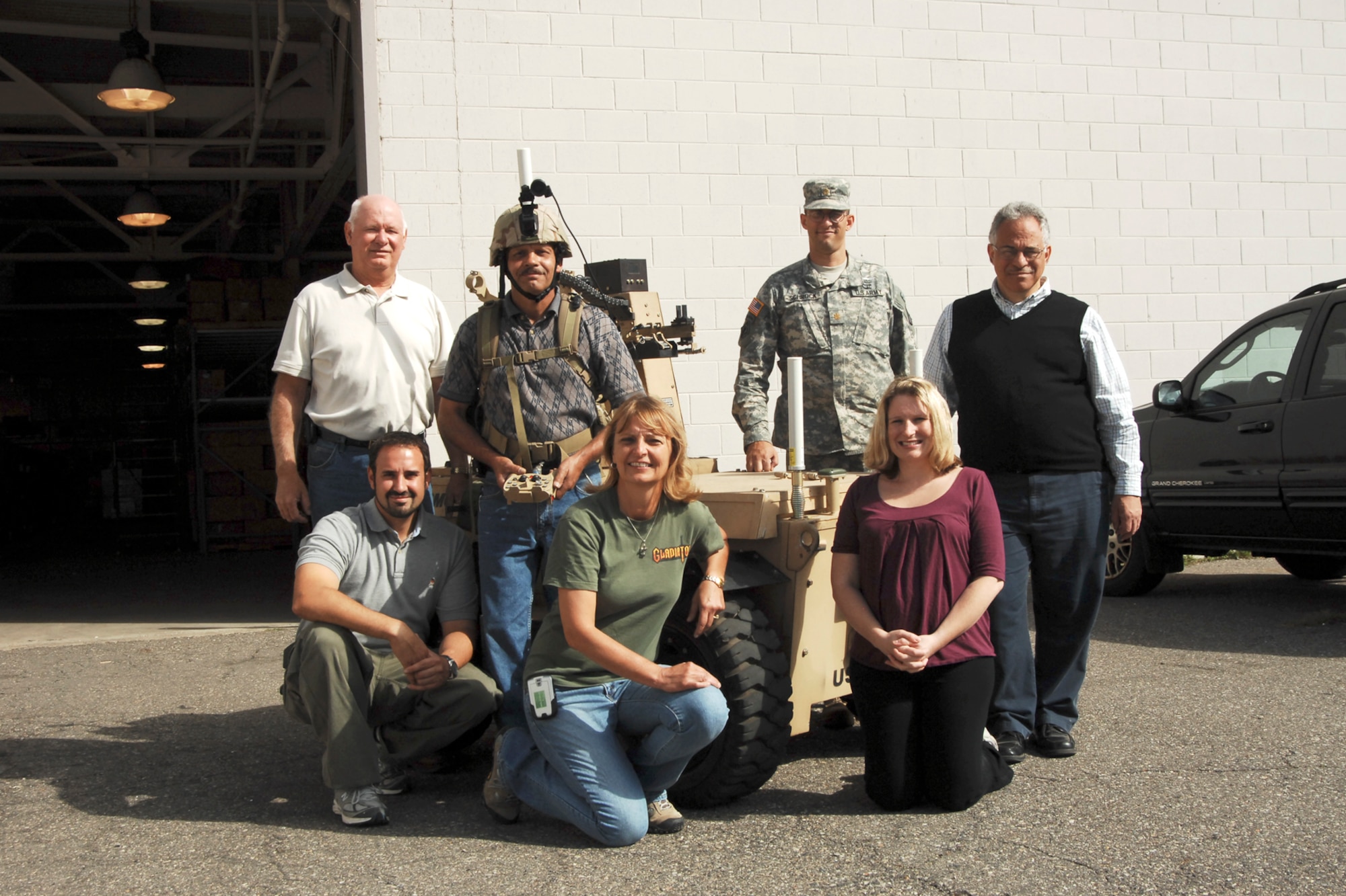 Personnel involved with the testing of the IbisTek wireless data link radio at Selfridge Air National Guard Base, Mich., last week, gather around the Gladiator robot.  The unique joint nature of the base in the northern suburbs of the metro-Detroit area, brought together many parts of the Army and the Air Force to create a cost-saving test environment for the radio which promised increased range for use on an Unmanned Ground Vehicle.  In this photo (left to right, starting back row), Mr. Greg Hudson, robot technician; JS RPO; Mr. Randall Ponder, engineering technician, JS RPO; the Gladiator Robot; Major Kevin Schrock, assistant manager, product APM maneuver system; Mr. Samuel Pirrone, radio frequency engineer; (front row) Mr. Robert Rappold, engineering team lead, Small Unmanned Ground Vehicles; Ms. Valerie Bolhouse, systems engineer; and Ms. Shanna Render, TARDEC Gladiator project manager.