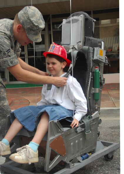 SHAW AIR FORCE BASE, S.C. -- Airman 1st Class Chris Demetres straps 7-year-old Hannah Bleymaier, daughter of Col. Steven Bleymaier, 20th Maintenance Group commander, into an ejection seat at the Sumter-Shaw Streetfest Sept. 25. Shaw's 20th Maintenance Group brought displays to the streetfest so families could see the work maintainers do on Shaw's flightline. (U.S. Air Force photo/2nd Lt. Emily Chilson)