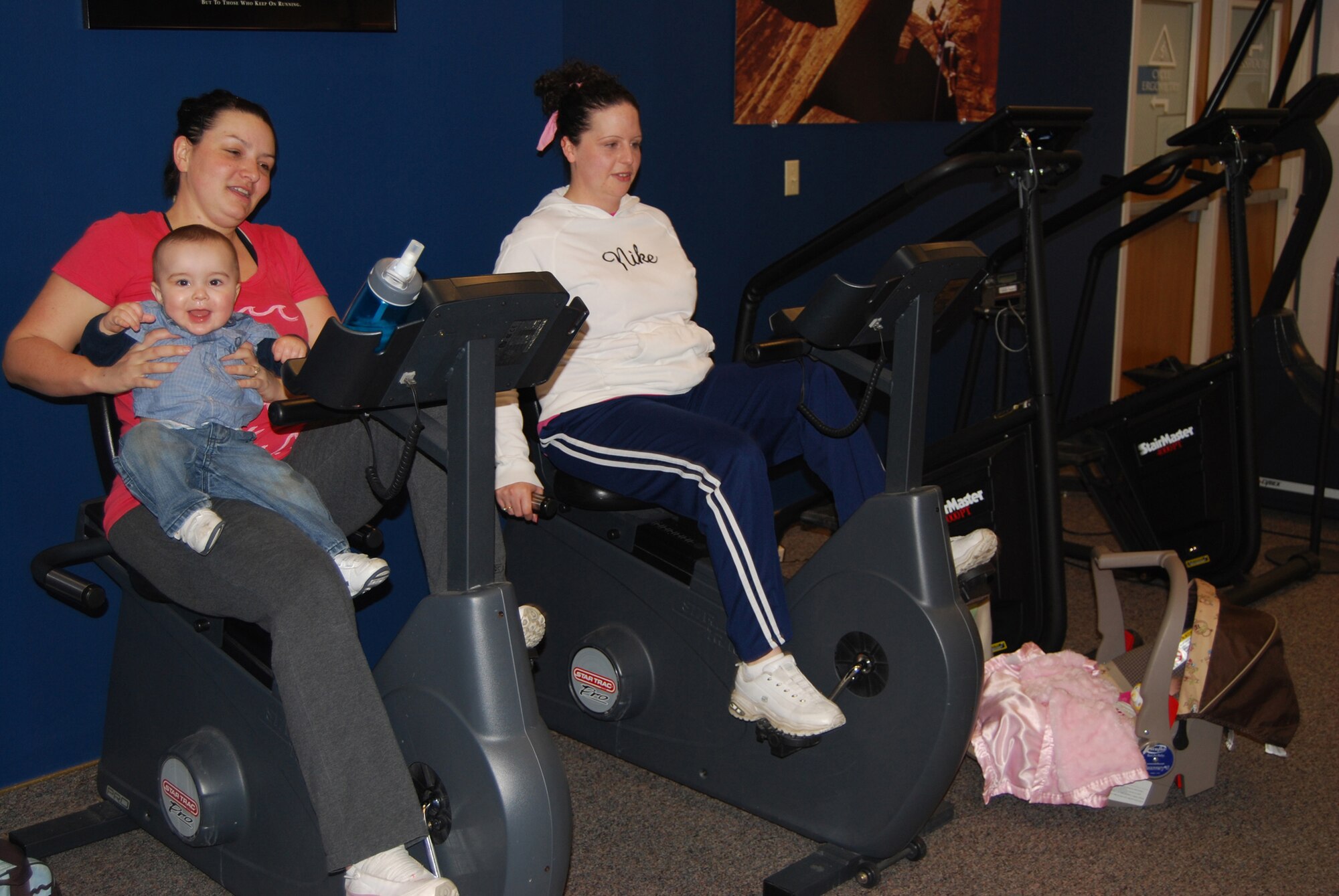 Jennifer Schoenbein, with son Tristan, 10 months, and Eliese Sago work out at the HAWC facility with their children March 30. The Health and Wellness Center also has a children’s area linked by video where parents can observe their children while they work out via monitors around the exercise area. (U.S. Air Force photo/Airman 1st Class Kristina Overton)
