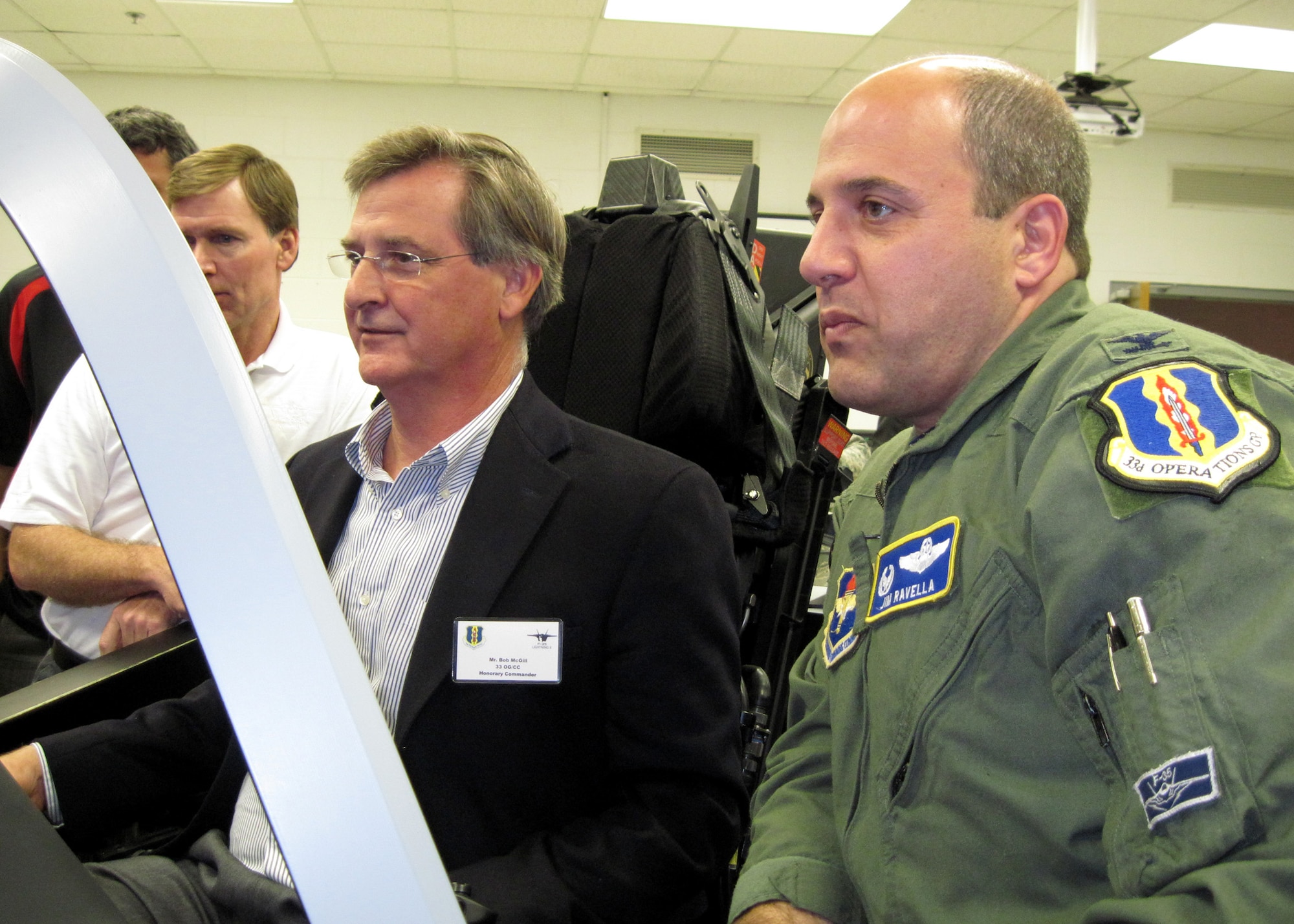 Col. James Ravella (right), 33rd Fighter Wing Operations Group commander, looks on as Bob McGill (center)“flies” the F-35 cockpit demonstrator. Mr. McGill is the new honorary commander for the 33rd OG and joined eight other new honorary commanders in the induction ceremony April 9. (Air Force Photo/Ashley M. Wright)