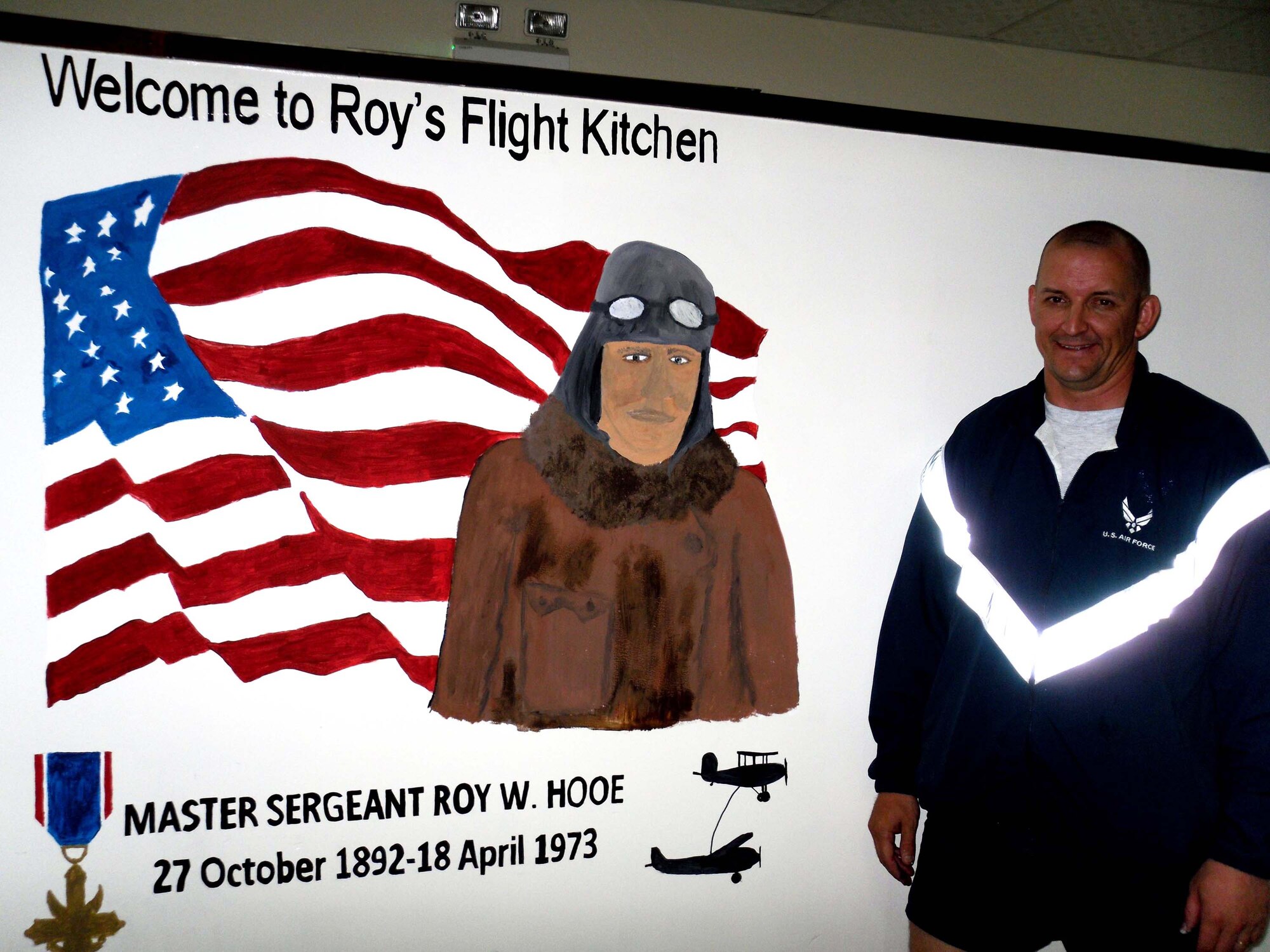 Master Sgt. Scott Sturkol, superintendent of 380th Air Expeditionary Wing Public Affairs at a non-disclosed base in Southwest Asia, stands next to a mural he helped design and paint April 18, 2010 at Roy's Flight Kitchen in the operations area for the wing honoring Master Sgt. Roy Hooe for whom the flight kitchen is named after. Sergeant Hooe is best known for his support of the first air refueling mission with the "Question Mark." Sergeant Sturkol is deployed from the Headquarters Air Mobility Command Public Affairs at Scott Air Force Base, Ill., and his hometown is Wakefield, Mich. (U.S. Air Force Photo/Master Sgt. Scott T. Sturkol/Released)