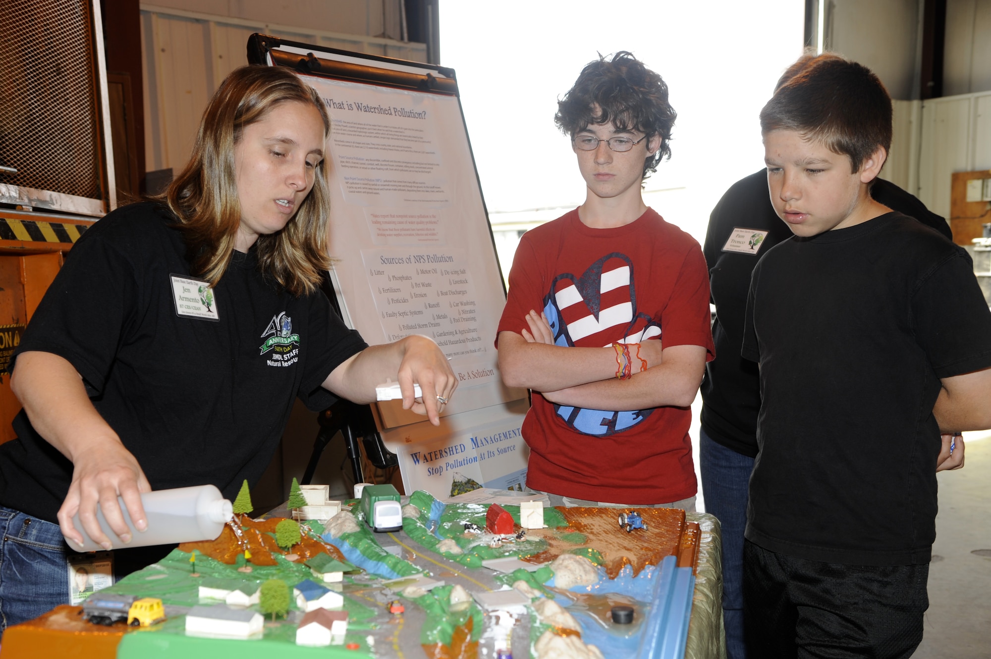 Jennifer Armento, 87th Civil Engineering Natural Resource Ecologist, uses a tabletop model to demonstrate how pollution from streets, housing areas and businesses get washed down into the local watershed and contaminates streams and other water sources during an Earth Day event April 21 at Joint Base McGuire-Dix-Lakehurst. Tyler Ferson, son of Jennifer and Army Lt. Col. Victor Ferson, 244th Aviation Brigade, and Andrew Lara, son of Rebecca and Army Maj. Erik Lara, 244th Aviation Brigade, watched and asked questions during the demonstration. The watershed is the area of land where all water that is under or drains off it goes into the same place. (U.S. Air Force photo/Carlos Cintron)