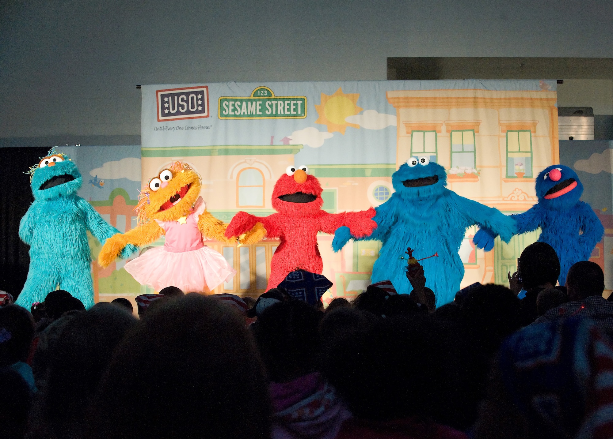 (Right to left) Rosita, Zoe, Elmo, Cookie Monster and Grover hold hands and sign during a Sesame Street live show hosted by the United Service Organization April 29 at the Youth Center. They got the audience involved at the end of the show, asking the children to get up and dance with them, as well as offering hugs and goodbyes as they left the stage. (U.S. Air Force photo/Brianne Zimny)