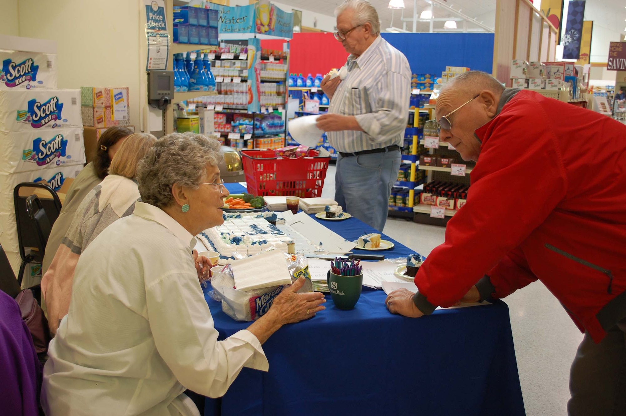 Montana Retiree Council member Vivian Peterson explains to retireeRon Sperandia, what other activities are available during the day when he stopped in to the commissary April 24. The Council members also manned welcome tables at the base exchange, clinic and Elkhorn Dining facility. Enjoying a piece of cake in the background is retiree Edward Olson. (U.S. Air Force photo/Valerie Mullett)