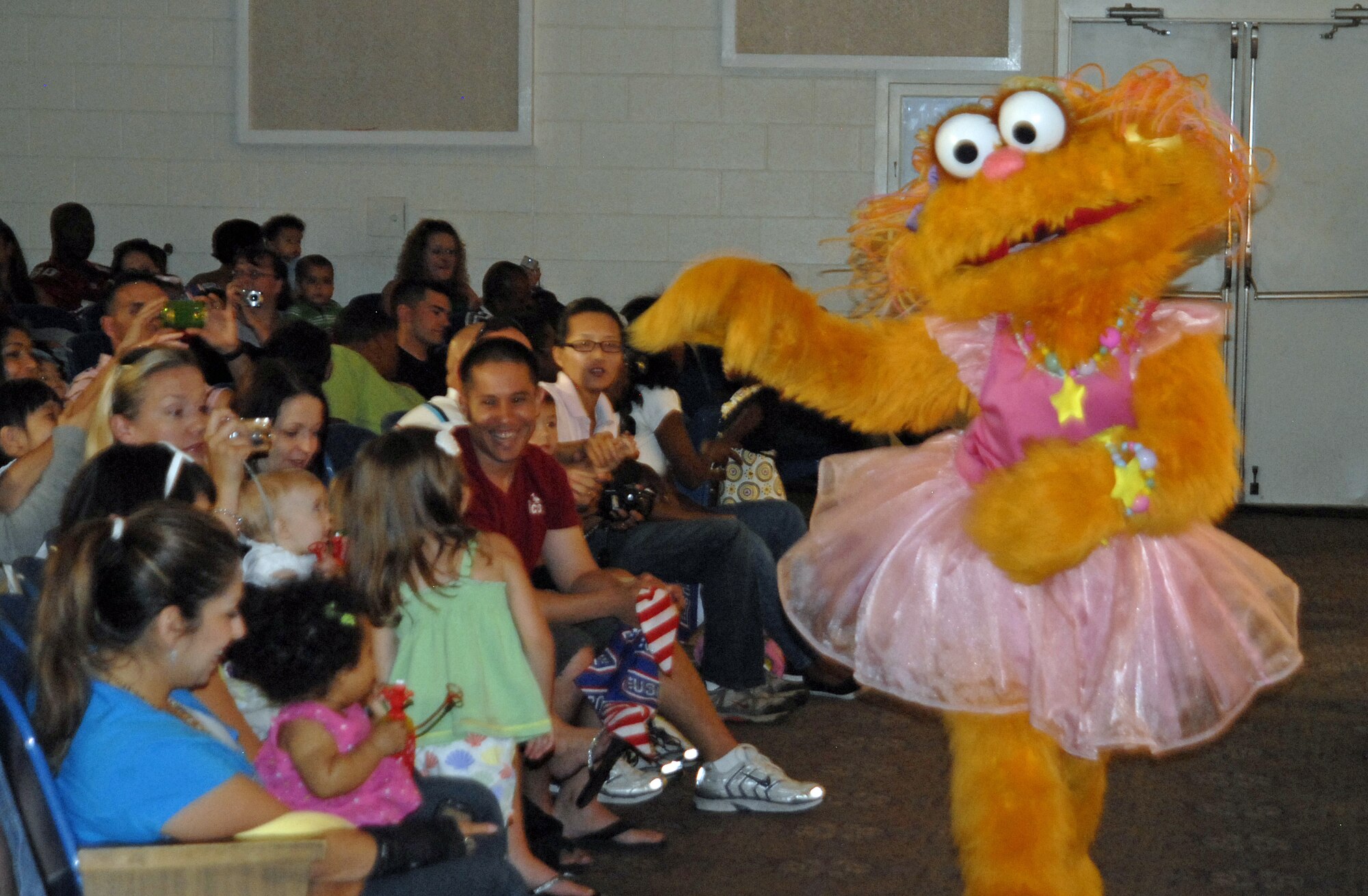 Parents and children watch and take pictures as Zoe, one of Elmo’s Sesame Street friends, takes time out of the performance to say ‘hello’ to audience members. The Sesame Street/USO Experience for Military Families is a free traveling USO show based on Sesame Workshop’s award winning “Talk, Listen, Connect” initiative. (U.S. Air Force Photo/Airman 1st class Jessica Hines)