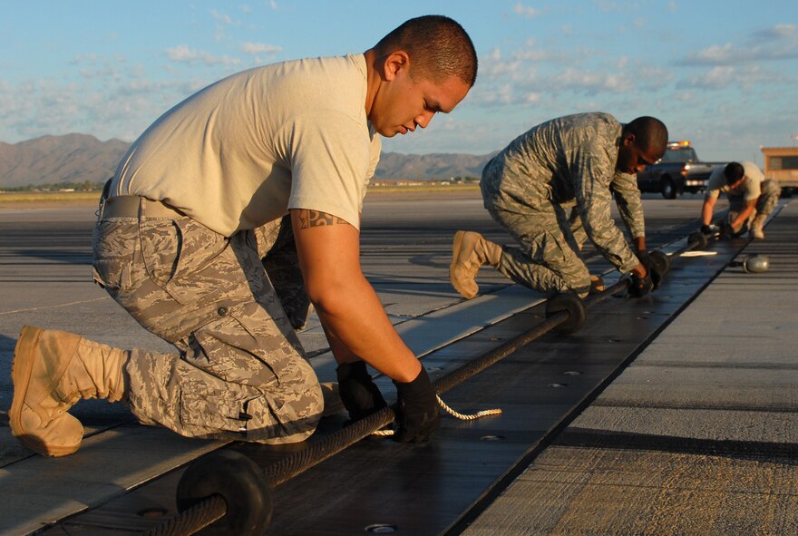 Senior Airman Justin Tayamen, Airman 1st Class Brian Thorne, and Senior Airman Gabe Ramirez, 56th Civil Engineer Squadron electrical power production journeyman, tie down the cable to barrier arresting kit 9 while setting it in service August 10th on the run way at Luke Air Force Base, Arizona. (U.S. Air Force photo/Airman 1st Class Sandra Welch)