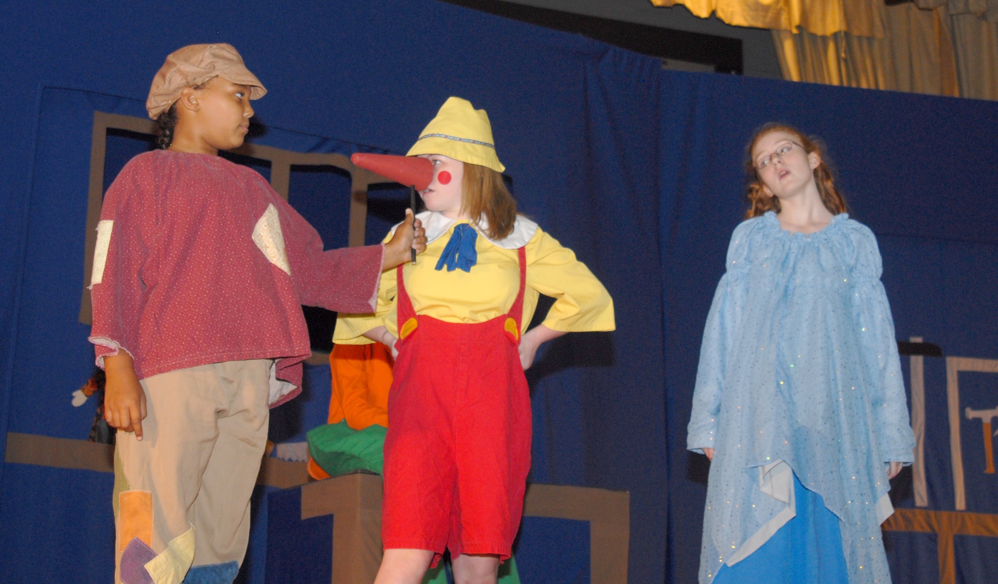 An Urchin, Alexia Goodman, holds Pinocchio's growing nose (played by Bethany Brown) while Blue Fairy, Isabelle Ingalls, tells Pinocchio that with each lie told, his nose will grow longer during the Aug. 21 show here. Auditions for Pinocchio were held at the beginning of the week casting 35 youths in grades K-12 to their roles. They rehearsed throughout the week, learning lines, songs and choreography performed two shows as an ensemble. (U.S. Air Force photo/Connie Hempel)
