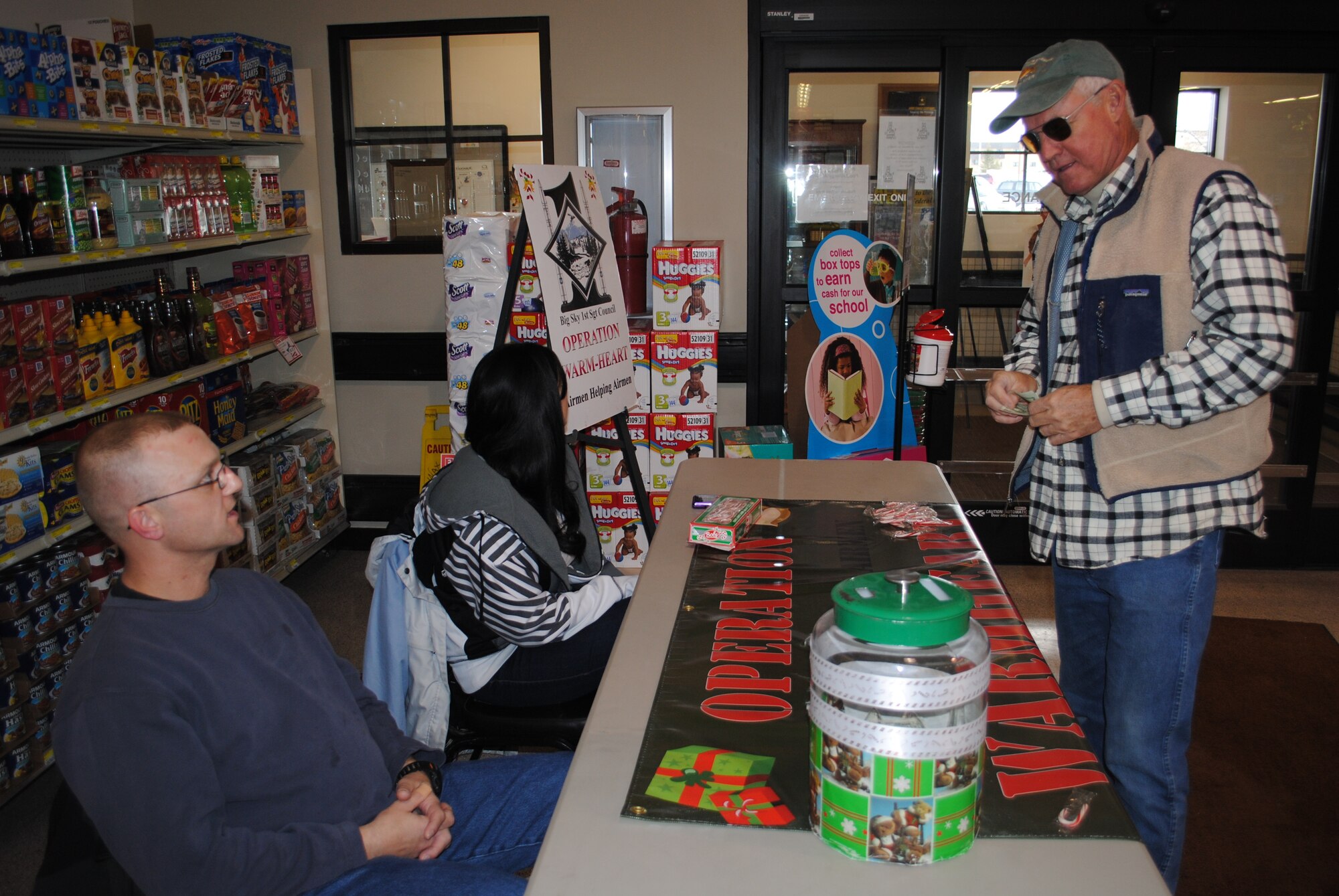 Master Sgt. Todd Smith, 341st Security Support Squadron first sergeant, and Airman Alexis Simpson, 341st Missile Security Forces Squadron member, look on as Mr. Pat Pauli donates to Operation Warm Heart during a visit to the commissary Nov. 30. OWH is a program managed by Team Malmstrom’s First Sergeants Council geared toward helping Airmen and their families undergoing financial hardships by providing them with grants to support them in their time of need. The program will continue at the base commissary until Dec. 19. (U.S. Air Force photo/Airman 1st Class Kristina Overton)