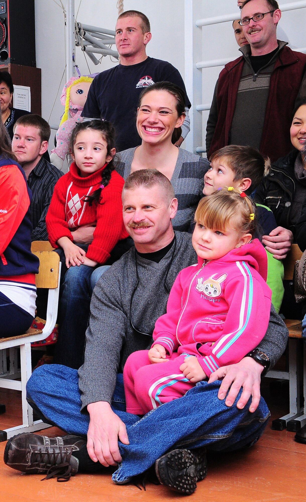 TRANSIT CENTER AT MANAS, Kyrgyzstan - Senior Master Sgt. Darrin Jetton, 376th Expeditionary Operations Group superintendent and Staff Sgt. Trish Arena, 22nd Expeditionary Aircraft Refueling Squadron aircrew flight equipment, sit with children as a local school performs for the New England Patriot Cheerleaders during their visit as part of an Armed Forces Entertainment tour Dec. 23.  The cheerleaders took a cultural tour of Bishkek, performed for deployed military members and toured the Transit Center at Manas. (U.S. Air Force photo/Staff Sgt. Stacy Moless)
