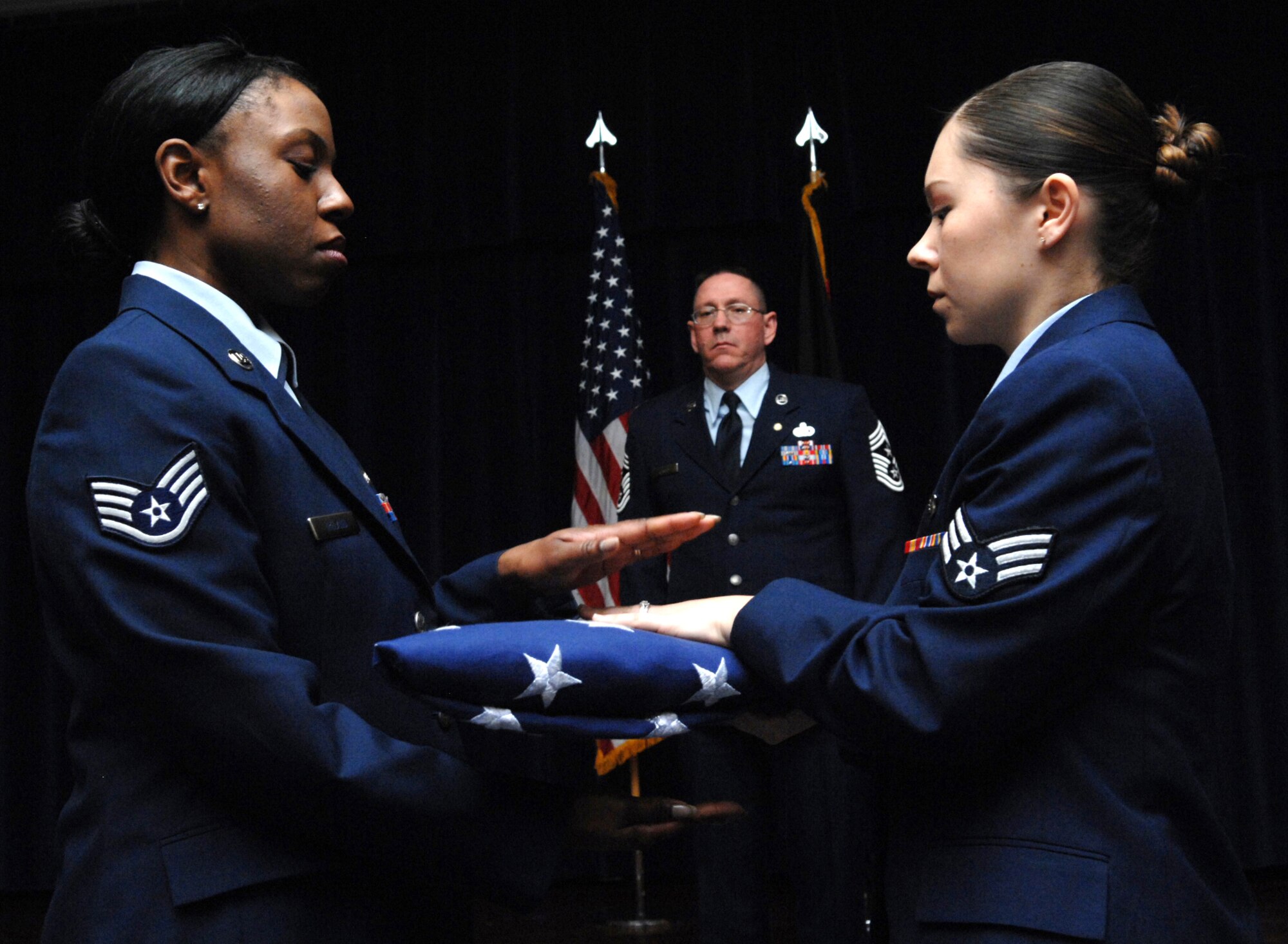 SPANGDAHLEM AIR BASE, Germany -- Staff Sgt. Marcie Ireland, left, 52nd Force Support Squadron lodging supervisor, takes a folded American flag from Senior Airman Tina Navarrette, 52nd Medical Operations Squadron aerospace medical technician, as retired Chief Master Sgt. Bruce Kenney, former 52nd Fighter Wing command chief, watches during the "Progression of Ranks" portion of his retirement ceremony Feb. 11 at Club Eifel. Chief Kenney served in the Air Force for 30 years. (U.S. Air Force photo/Airman 1st Class Nick Wilson)