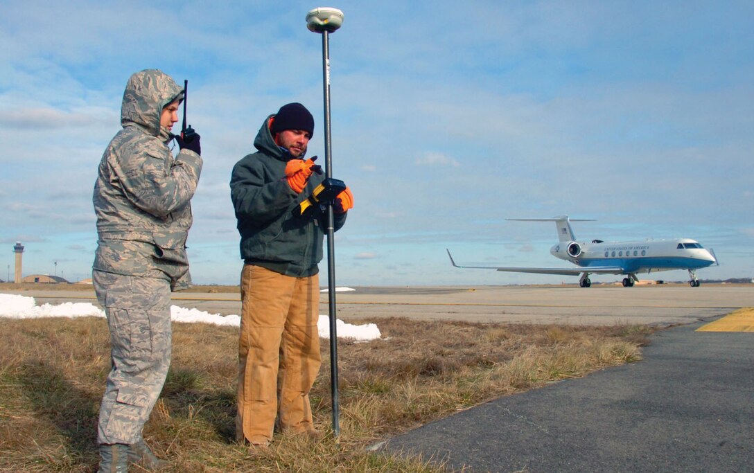 Airman 1st Class Amber Hernandez, 316th Force Support Squadron force management representative, escorts Jason Hall, contractor, as he takes land elevation readings during a survey of the runway near the Whiskey Taxiway, while a 89th Airlift Squadron C-20 aircraft prepares to take off on the flightline Tuesday. The 316th Civil Engineer Squadron is overseeing several contract agencies currently conducting surveys, taking soil samples from borings and managing water drainage pipes under the flightline for the purpose of overall future ramp improvements. (U.S. Air Force photo/ Bobby Jones)