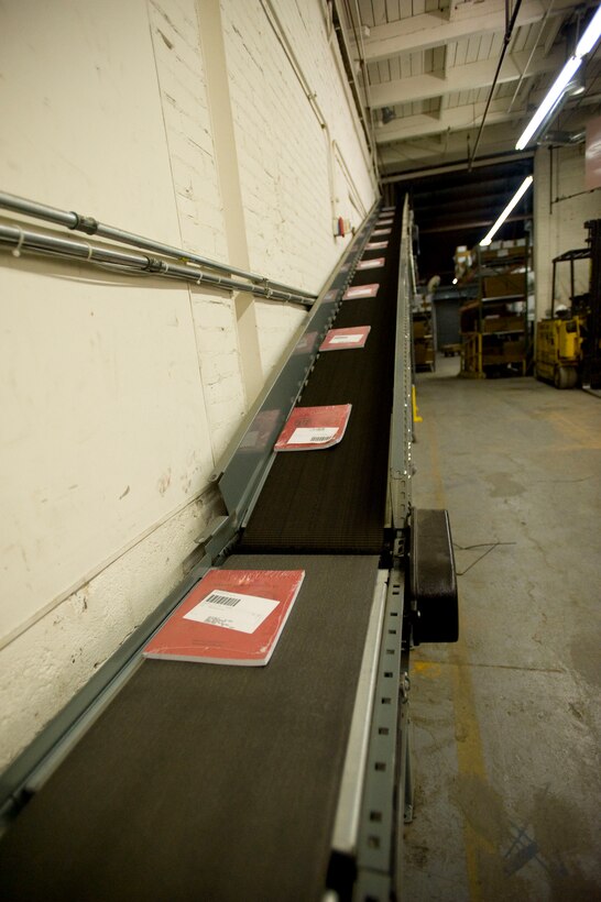 The 359 ft. conveyor belt winds ascends high into the Marine Corps Institute warehouse, allowing clearance for the forklift to operate underneath. The belt then descends into the postal section, where automated rollers sort books into mailing bags for shipment.
