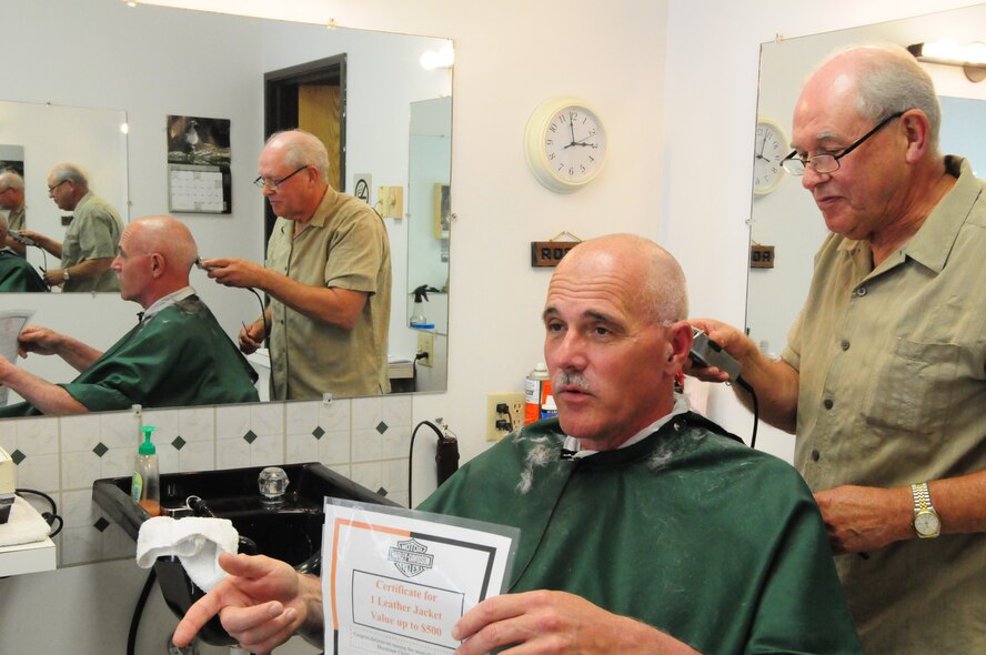 Retired Happy Hooligan Roger Dissmore, of North University Barbers, Fargo, N.D. gives a closely-cut haircut to Chief Master Sgt. David Harmon, the 119th Wing command chief, June 29.  Harmon is getting the haircut in agreement to a challenge met by 119th Wing members Master Sgt. Brian Rook and Senior Master Sgt. Wade Swenson.  Harmon challenged Rook and Swenson to raise donations for the Fargo chapter of the Harley owners' group Miracle Ride.  The Miracle Ride money goes to the Meritcare Children's Hospital in Fargo. Chief Master Sgt. Harmon is sitting in the barber chair and holding a flyer advertising the Miracle Ride, for which he raised $3,019 with the help of Rook and Swenson. 