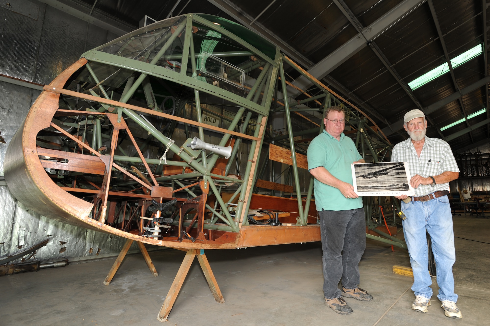 WHITEMAN AIR FORCE BASE, Mo. - Frank McKinley, of the Whiteman Heritage Foundation, and Robert Rainey, Waco glider restoration project volunteer, hold an artist's rendition of a Waco glider in flight while standing in front of the original glider they are restoring, July 6.   In 1942, U.S. Army Air Corps officials selected the site of present-day Whiteman AFB to be the home of Sedalia Army Air Field, a training base for Waco glider pilots. (U.S. Air Force photo/ Senior Airman Carlin Leslie)(Released)
