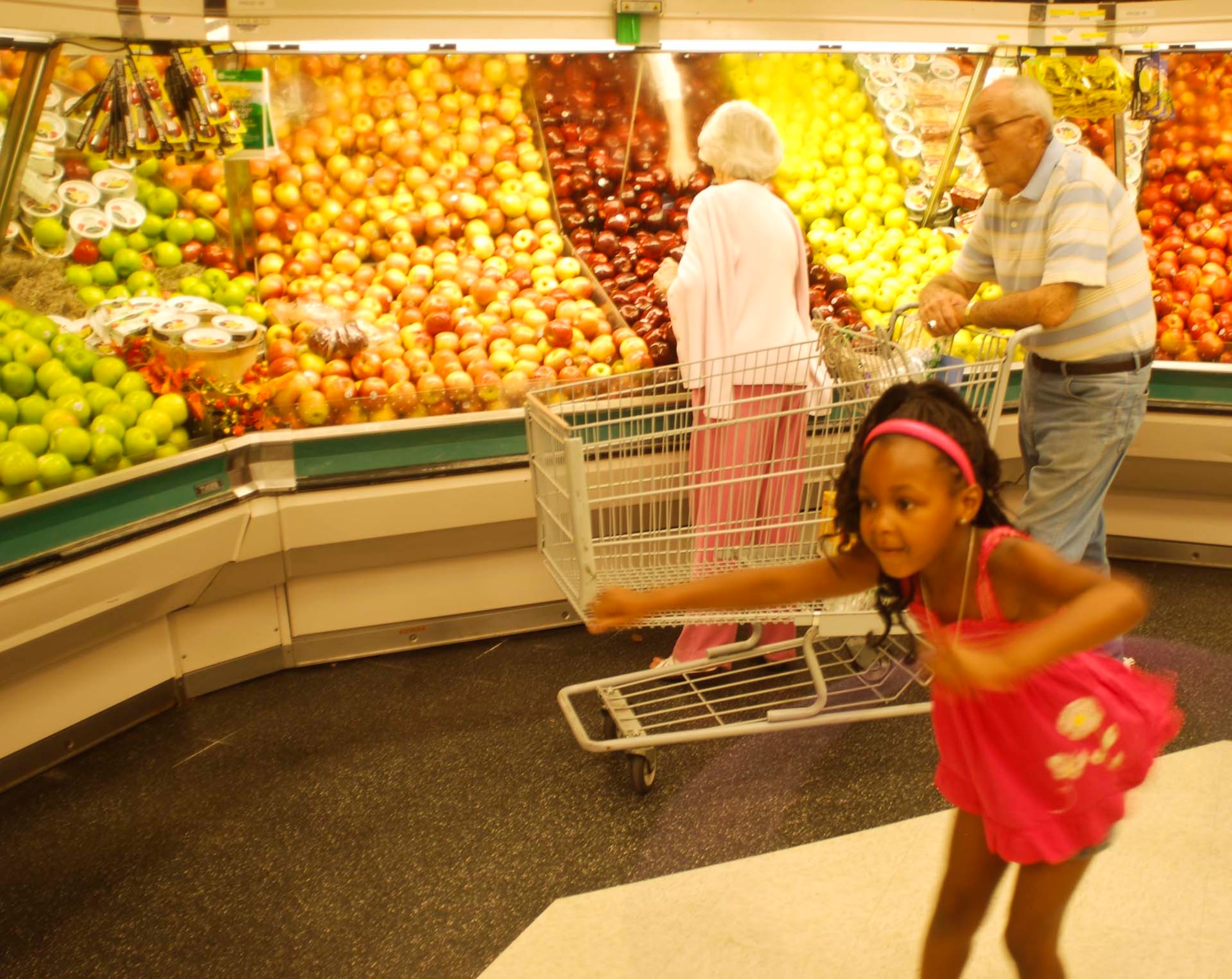 Journee, the granddaughter of March Commissary management specialist, Ponsiette Lynch, shows off her hula-hooping skills as commissary shoppers behind her select apples from the refrigerated display case.  (U.S. Air Force photo by Megan Just)