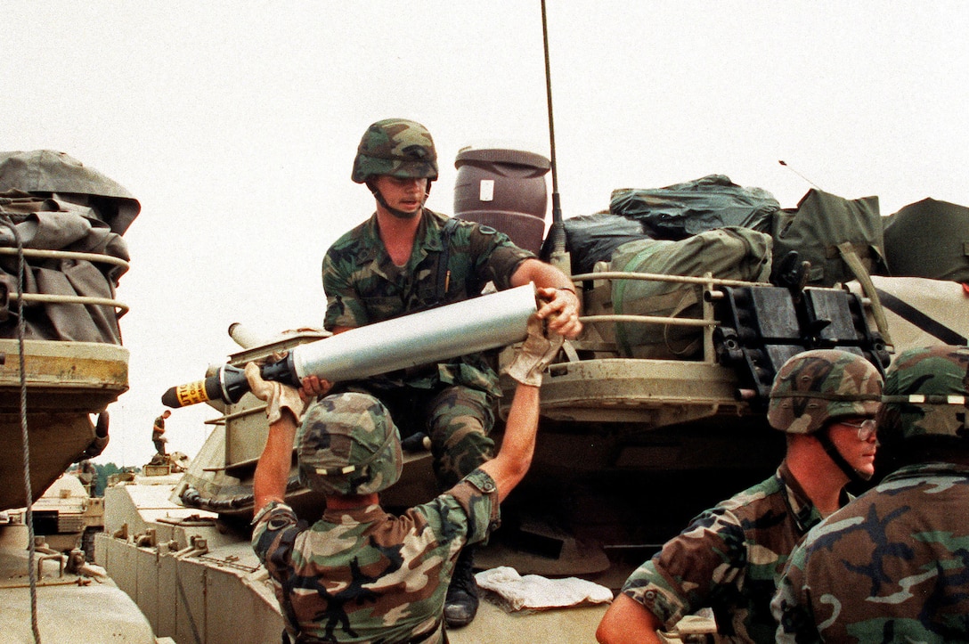 U.S. Army Pfc. Edward Bessette hands a 105mm round up to Spec. Scott Arnold as soldiers of 1st Battalion, 64th Armor, 24th Infantry Division (Mechanized) load ammunition aboard their M-1 Abrams tanks prior to deployment to Saudi Arabia for Operation Desert Shield.