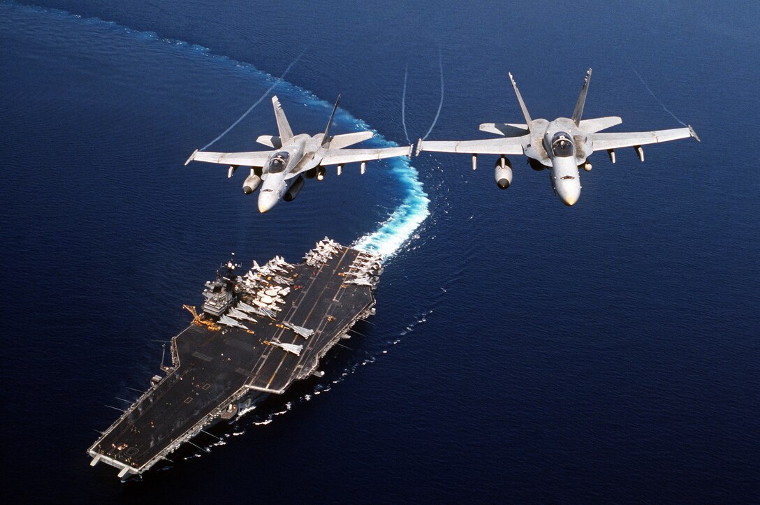 Two F/A-18C Hornet aircraft of Strike Fighter Squadron 74 fly above the Forrestal class, aircraft carrier USS Saratoga making a hard turn to starboard during Desert Shield.  