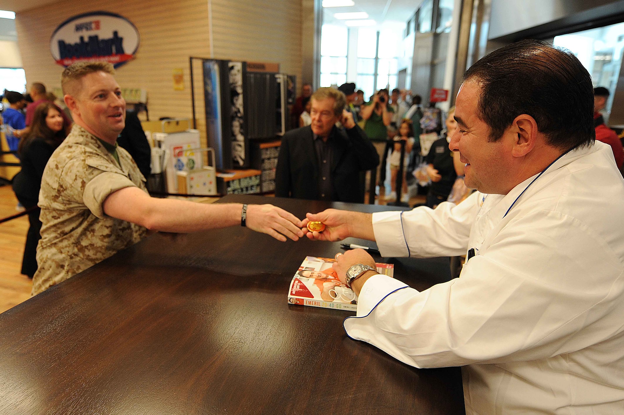 U.S. Marine Corps Gunnery Sgt. Ben Coleman, Wounded Warrior Center representative, presents Chef Emeril Lagasse with a unit coin during a book signing at the Kaiserslautern Military Community Center, Ramstein Air Base, Germany, May 29, 2010. Celebrity Chef Emeril Lagasse visited the KMCC to promote his new book along with providing a book signing and a full production cooking exhibition copies of his new book and performing an hour long cooking demonstration for over a thousand visitors. (U.S. Air Force photo by Staff Sgt. Stephen J. Otero)