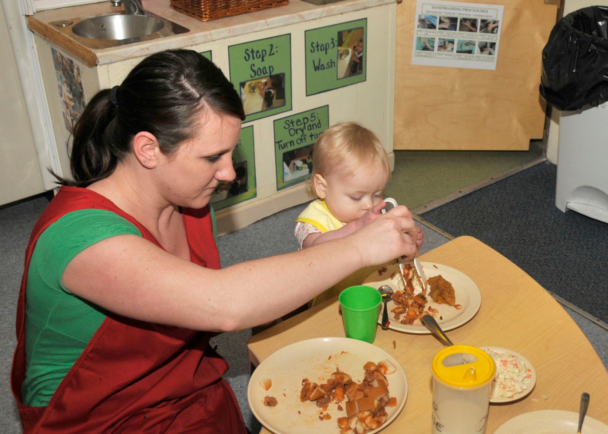 Dana Fraley, a caregiver at Child Development Center East, helps one-year-old Caitlyn Mabie, daughter of Kristy Mabie, an Education and Training Technician at CDC East, serve herself lunch Feb. 26 at RAF Lakenheath.  Caregivers encourage self-help and build fine motor skills by practicing family-style dining for all meals served at the facility in accordance with the criteria outlined by the National Association for the Education of Young Children. (U.S. Air Force photo/Capt. Alysia Harvey)