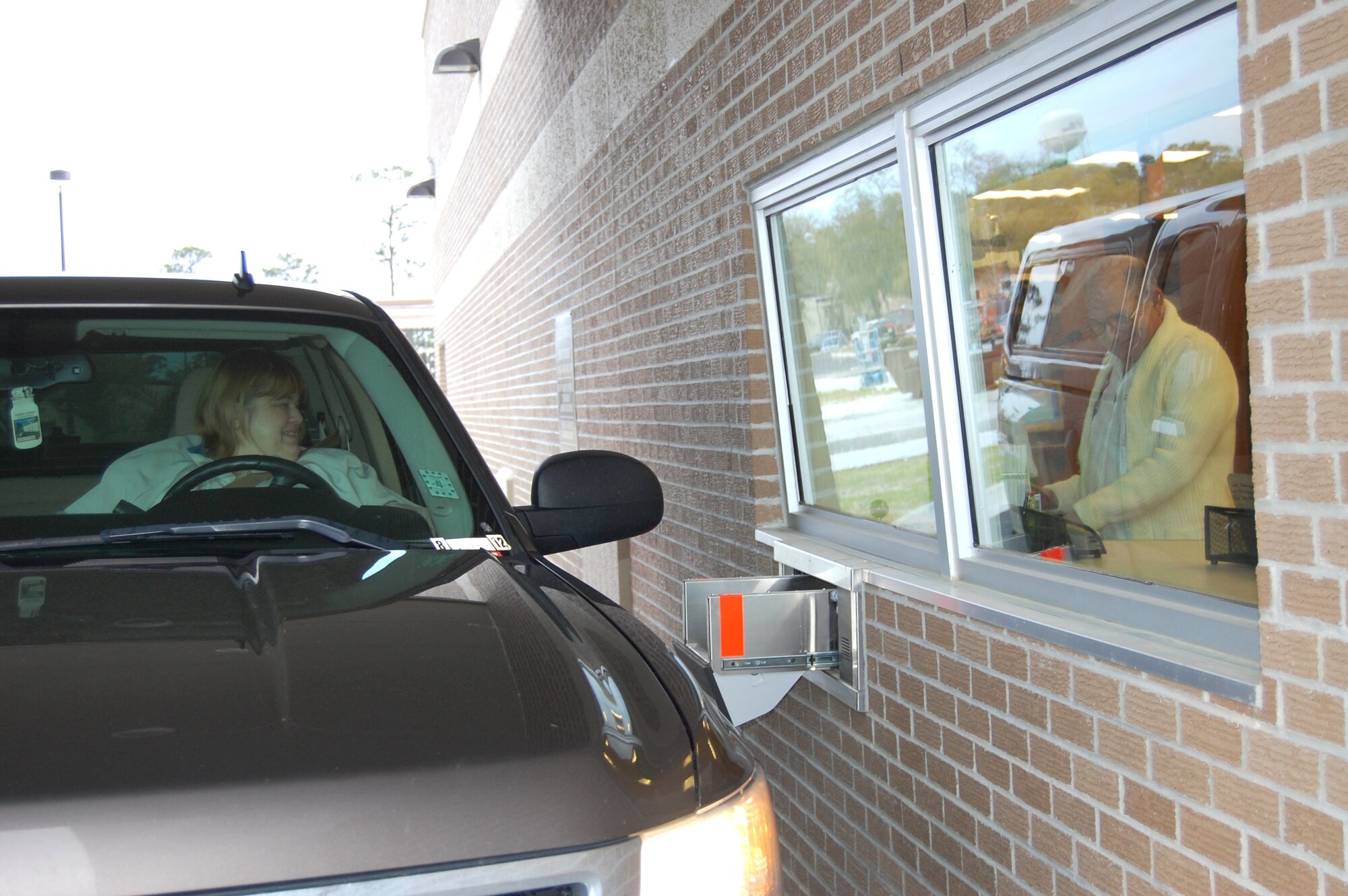 Bonnie Forgie is the first drive-up customer at the new satellite pharmacy that opened March 22 as 10-year volunteer Richard Dunlap mans the window.  The new facility is about twice as large as the trailer it replaces and slightly larger than the satellite pharmacy destroyed by Hurricane Katrina in 2005.  The staff includes eight pharmacists and 19 technicians serving between 750-1,000 patients and dispensing between 1,400-2,000 prescriptions every duty day.  Hours are 8 a.m. to 5 p.m. weekdays.  The double drive-through window is open until 5:3 p.m. weekdays and 8 a.m. to 2 p.m. Saturdays for pickup of completed prescriptions and refills only.  (U.S. Air Force photo by Steve Pivnick)