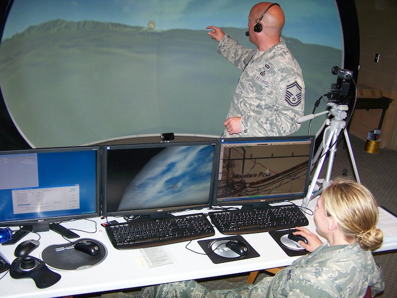 A trainee (standing) calls in an airstrike to “destroy” an enemy tank during joint combat air support training on the Joint Terminal Attack Controller virtual training dome, while another trainee (seated) enters information enabling the simulated aircraft to react as intended. 
(Air Force image)