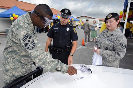 Chief Master Sgt. Juan Lewis, 502nd Air Base Wing command chief, reaches for a pen while wearing glasses that simulate the visual impairment caused by alcohol or other drugs. Officer Ruben Morales of the Converse Police Department and Staff Sgt. Marisela Gonzales, 902nd Security Forces Squadron, evaluate his performance during safety day to kick off 101 Critical Days of Summer. (U.S. Air Force photo/Steve Thurow)