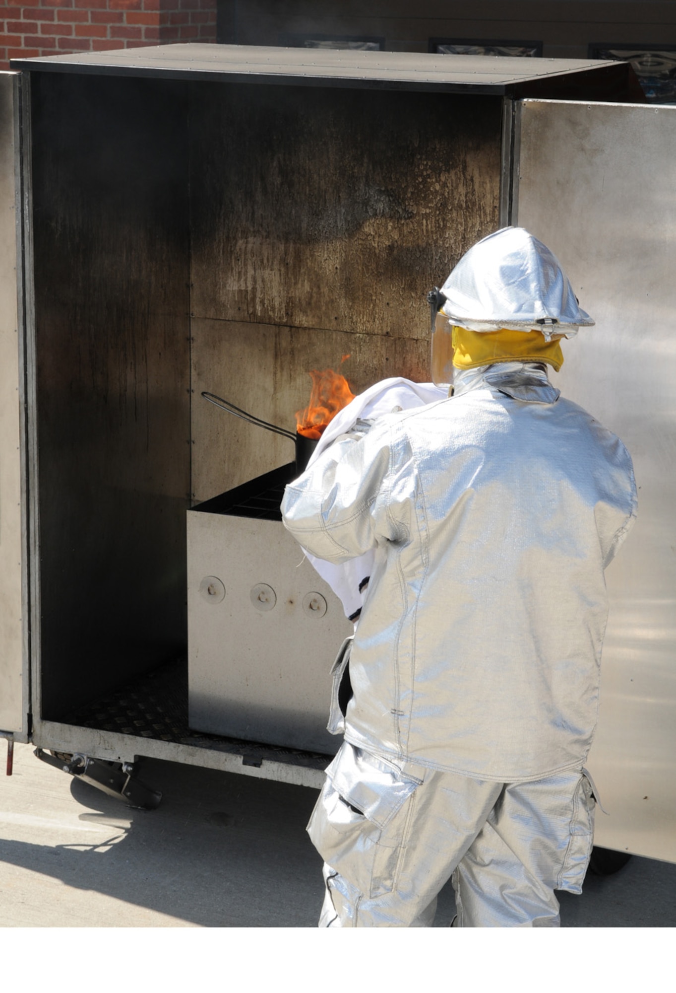 Firefighter Mark Peskett, Defence Fire Service firefighter, 100th Civil Engineer Squadron, covers the grease pan fire with a damp towel to extinguish it. He then removes the towel and, from a distance, pours water onto the pan to show what can happen if someone did pour water onto the fire instead of putting it out properly. (U.S. Air Force photo/Karen Abeyasekere)