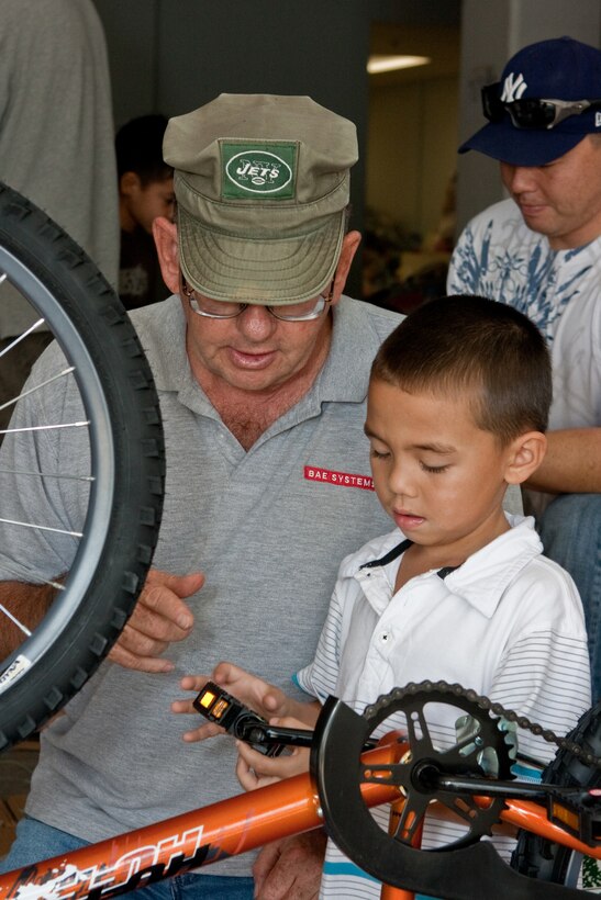 Frank McLaughlin III and his grandson, Frank McLaughlin V work together assembling one of the bicycles to be donated while Frank McLaughlin IV works on another bike behind them Nov. 20 during BAE Systems' bike build.  The eldest McLaughlin is a BAE Systems employee and he brought three generations of his family to help out with their annual "Bikes for Tykes" program, which has donated nearly 500 bikes to Hawaii's less fortunate children through the Marine Corps Reserve's Toys for Tots Hawaii program over the past five years. (Official U.S. Marine Corps photo by Chuck Little)