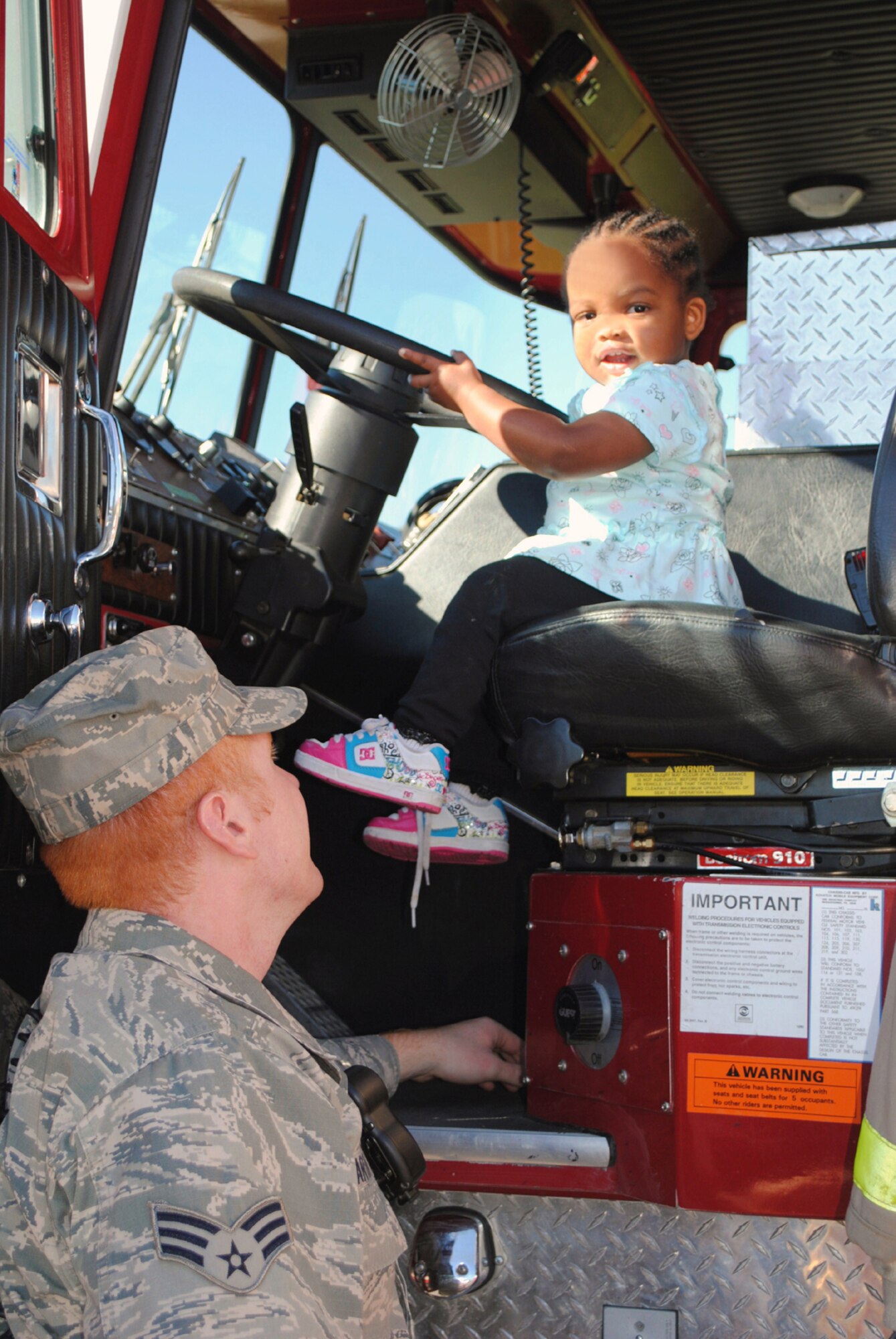Senior Airman Mark Martin, 341st Civil Engineer Squadron firefighter, assists Little Warrior Kay-Leigh, 1, as she takes the wheel of a fire engine. (U.S. Air Force photo/Airman 1st Class Kristina Overton)