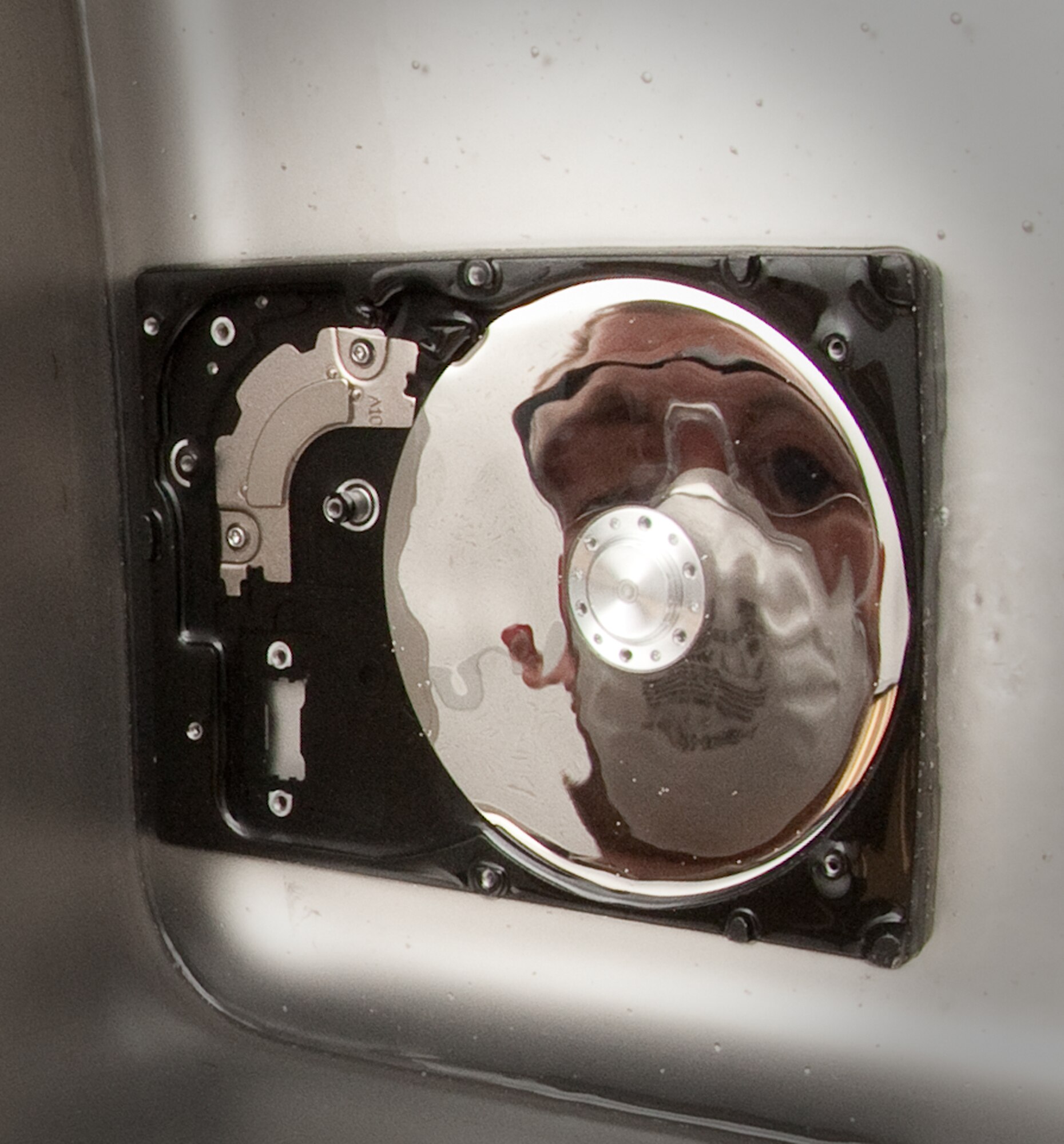 Scott Lalliss's reflection is seen on the platen disk of a hard drive being cleaned in a sonic cleaner with 91 percent alcohol solution at the Defense Computer Forensic Laboratory Sept. 9, 2010, in Linthicum, Md. The 9 percent of water in the solution develop bubbles that gently move dirt and other organic matter away from the disks that may contain valuable evidence in an investigation. Mr. Lalliss is a senior forensic technician. (U.S. Air Force photo/Lance Cheung)