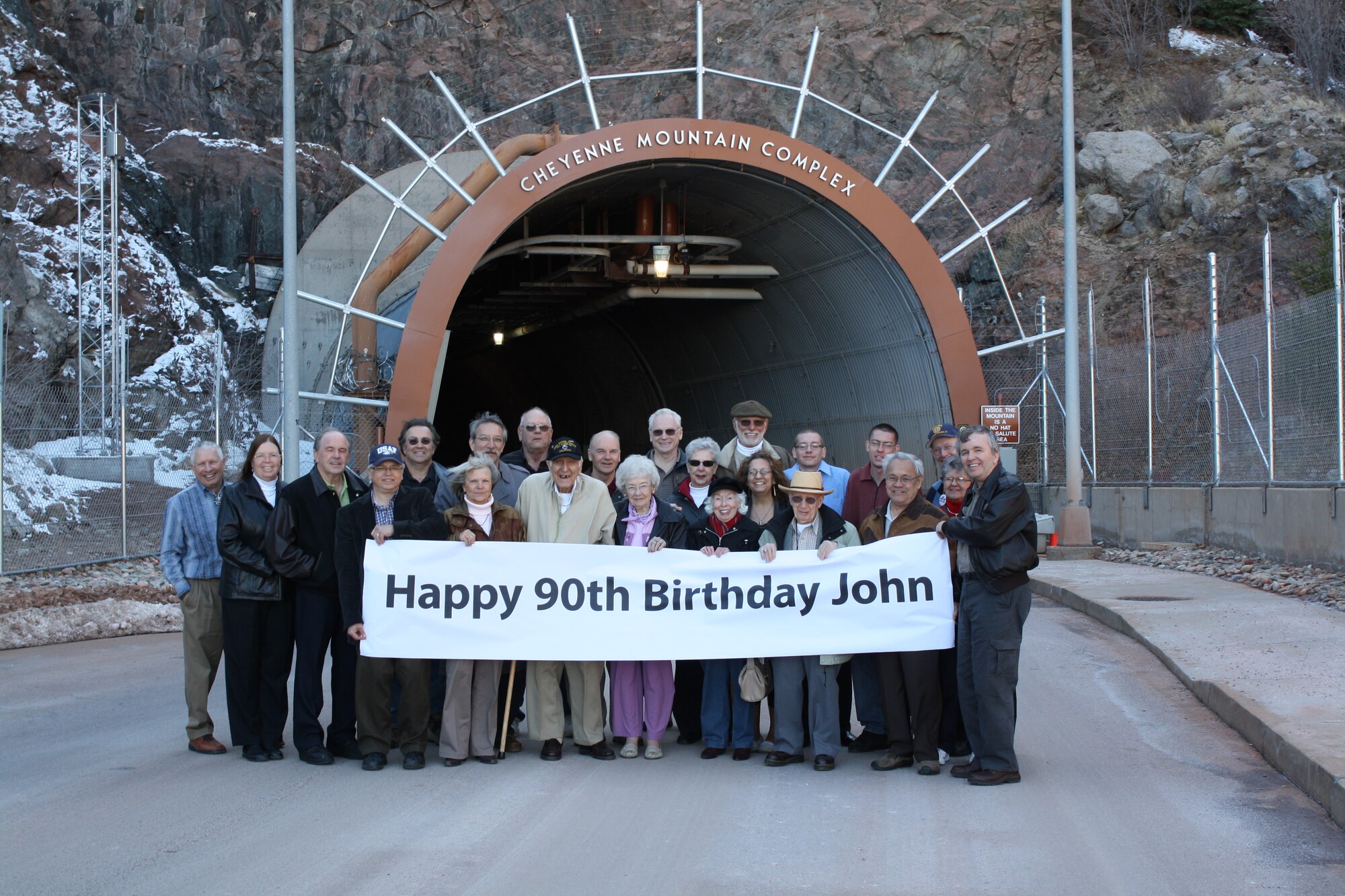 Retired U.S. Air Force Master Sgt. John Mutu (front row center with black ball cap), gathers with friends and family for a surprise 90th birthday April 4 on Cheyenne Mountain Air Force Station by 22 former colleagues and family members. The group, which worked together in the communication squadron, shared stories of the "good old days," toured the mountain and noted some of the remarkable changes to their former workstation. In 1979, carrying on family tradition, Bob Mutu (far left), Mr. Mutu’s son, went to work at CMAFS in the electric shop, where he still works as a DOD employee. Mr. Mutu had a long distinguished career in the U.S. Air Force, but said some of his fondest memories were of working in a special place like CMAFS. (U.S. Air Force photo/1st Lt. Daniel Taylor)