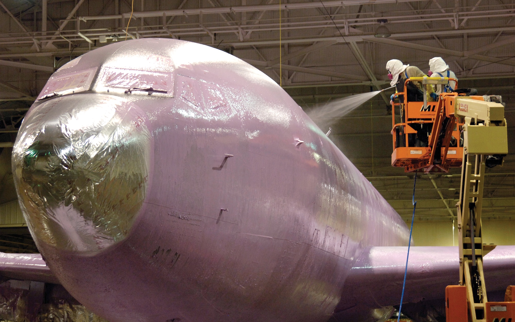 In the Tinker Air Force Base depainting facility of the 76th Aircraft Maintenance Group’s Service Section in Bldg. 2122,depainters use a lift to reach the top of a KC-135 Stratotanker with the pink aircraft chemical Plane Naked. Meticulous preparation and masking takes place before depainting, safeguarding areas not to be coated.  In less than an hour, the aircraft will be completely covered in the pink chemical as the process begins working over several hours. Standardizing preparation  methods is one way the unit has trimmed time off their production,  though they now work with crews nearly half the number as in  past years. ( U.S. Air Force photo by Margo Wright)