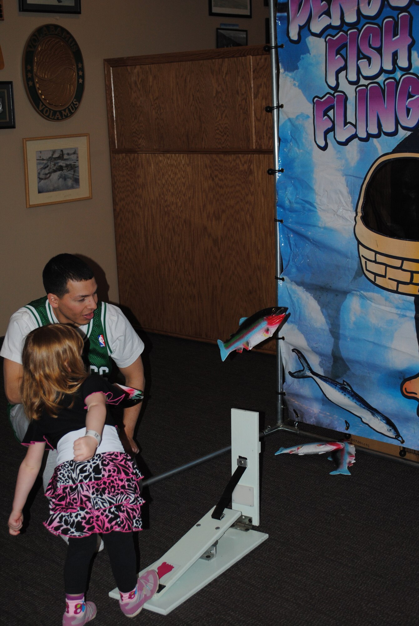 Shae Sweeney, 4, launches a fish at the 'Penguin Fish Fling' game while volunteer, Airman 1st Class Emmanuel Vargas, 341st Logistics Readiness Squadron vehicle operator, watches and cheers her on.  The Penguin Fish Fling was one of many games available for children to play at the carnival.  (U.S. Air Force photo/Airman Cortney Hansen)