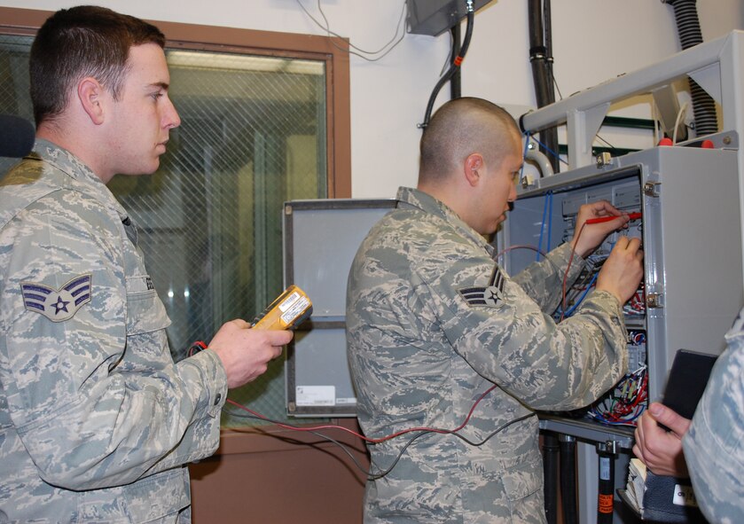 Senior Airman Alejandro Cedillos tests the voltage on a training environmental control system while Senior Airman Jeffrey Estes waits to get the reading. Staff Sgt. Kyle Babbert read instructions from the tecnhical order to the two periodic maintenance technicians with the 341st Missile Maintenance Squadron April 26 in the T-22 trainer. (U.S. Air Force photo/Valerie Mullett)