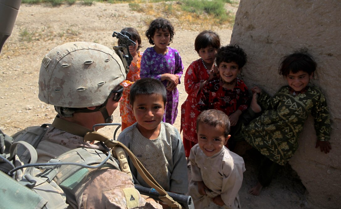 A Marine with Company L, 3rd Battalion, 4th Marine Regiment, bends down to greet local children in a small village outside his base. Children frequently come out and watch inquisitively at the Marines patrolling by, the girls dressed in brightly colored clothing, while boys generally wear clothing with little decoration.::r::::n::