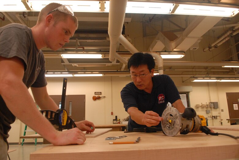 KUNSAN AIR BASE, Republic of Korea -- Mr. Chae-Yong Kim, a woodworker at the Arts and Crafts Center here, explains the router depth to Senior Airman Philip Bennett, 8th Maintenance Operations Squadron weapons standardization load crew member. Bennett is using the wood shop to build a dartboard for his squadron that will double as a bulletin board in his office. (U.S. Air Force photo/Master Sgt. Sonny Cohrs)