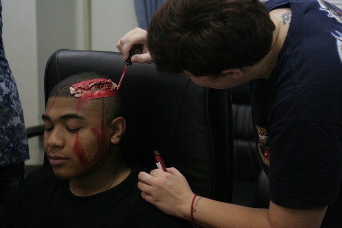 Lance Cpl. Jordan E. Payne, a mass casualty participant, sits with a severe head injury prosthetic on his head as Petty Officer 1st Class Katie A. Zirkle, Robert M. Casey Medical and Dental Clinic emergency medical technician, applies makeup before a mass casualty drill, which took place next to the station fire department here Aug 17. Participants were fitted with various prosthetics and makeup to simulate injuries and were placed near the Aircraft Rescue Firefighting downed aircraft training building.