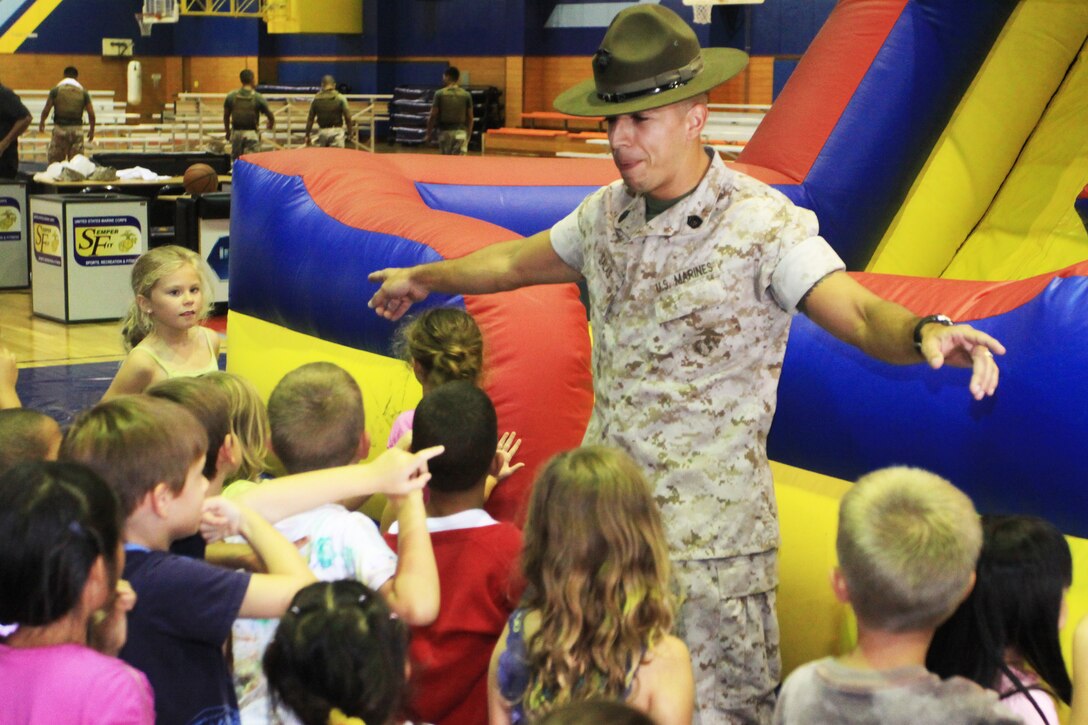 Gunnery Sgt. Fabio Salas, Marine Aviation Logistics Squadron 12 squadron gunnery sergeant, instructs a group of Matthew C. Perry first grade students to gather around before sending them through the bounce house obstacle course during the Johnny Wayne day at the IronWorks Gym sports courts here Aug. 23. During the event, students saw a Marine Corps Martial Arts Program demonstration, participated in sumo bashing and worked as a team to complete the obstable course.