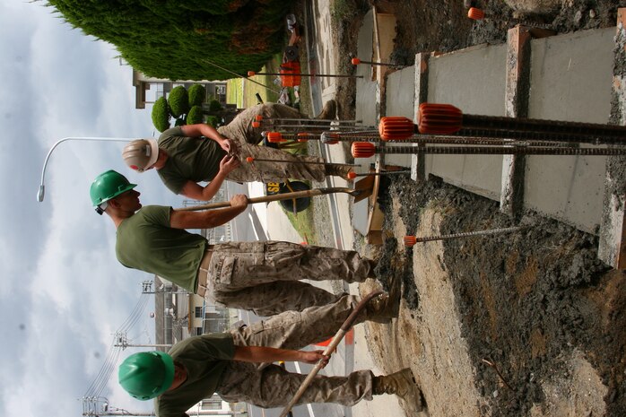 Lance Cpl. Adam Bailey (Left), Lance Cpl. David Waldbillig, both combat engineers, and Petty Officer 3rd Class William M. Svoboda, project manager, dig excess concrete and dirt away from the foundation of the new concrete wall at the Hornet’s Nest here Sept. 4. Excess dirt that makes its way into the concrete slab will create weak points because as the dirt slowly deteriorates, holes are left where the concrete needs to be making the whole wall weaker.