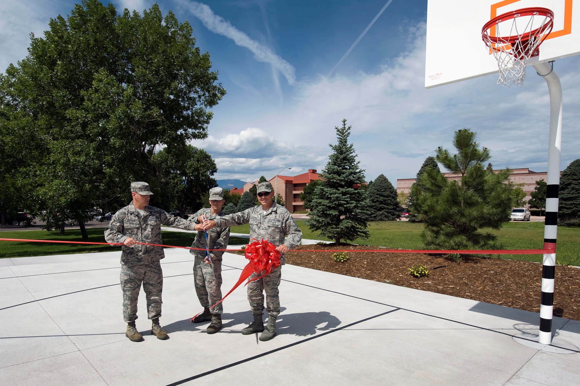 PETERSON AIR FORCE BASE, Colo. -- Chief Master Sgt. Thomas Trottier, 21st Space Wing command chief (left), and Chief Master Sgt. Randy LaCombe, 50th Space Wing command chief (right), assist Airman 1st Class Shaun Moon, 21st Dental Squadron dental laboratory apprentice, as he cuts the ribbon officially opening new basketball courts, Aug. 19, next to building 1164. The dorm council hosted a basketball tournament offering food and music, following the ribbon-cutting. The construction of the basketball courts was part of a $153,000 quality of life improvement project for Airmen who reside in Peterson dormitories. (U.S. Air Force photo/Craig Denton)