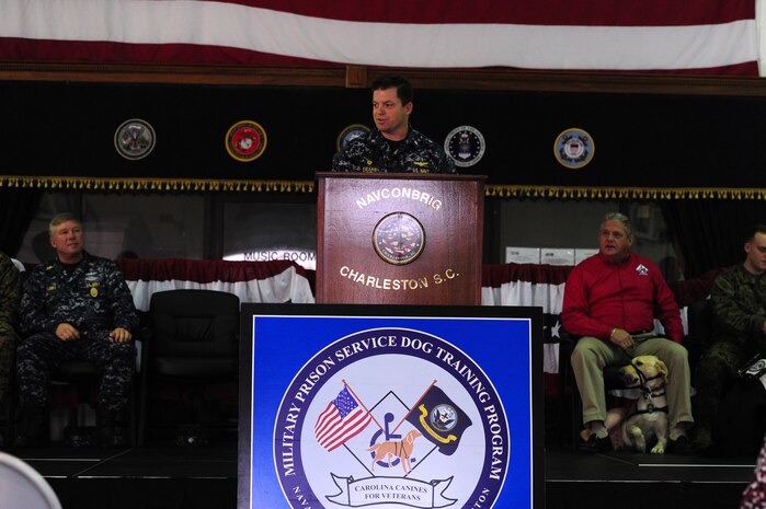Navy Cmdr. Raymond Drake gives opening remarks during a ceremony at the Naval Consolidated Brig Charleston Dec. 1.  During the ceremony, the NAVCON Brig, in partnership with Carolina Canines for Service, placed a prisoner-trained three and a half year-old black lab mix named Ruth with Marine Cpl. David Donchess, a wounded service member. Donchess deployed twice to Helmand Province, Afghanistan with the 1st Battalion, 6th Marine Regiment.  In March 2010, the seven-ton truck he was driving detonated an Improvised Explosive Device. He sustained numerous injuries and was awarded the Purple Heart. CCFS is a non-profit health and human services organization that trains service dogs for people with disabilities. Through this program, military prisoners are taught to train service dogs for veterans with disabilities. Since the program's inception, 14 wounded service members have received service dogs. Drake is the NAVCONBRIG commanding officer. (U.S. Air Force photo/ Staff Sgt. Nicole Mickle)  