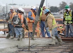 A construction worker guides a black 6-foot boom Dec. 9 as concrete flows onto the new Wilford Hall parking garage deck while the others use portable vibrators on their back to allow the mix to flow smoothly and spread evenly. The first placement of concrete marks the beginning of a 1,000-car parking garage for the ambulatory surgical center on Lackland Air Force Base, Texas. (Courtesy Photo/Will Ewing)

