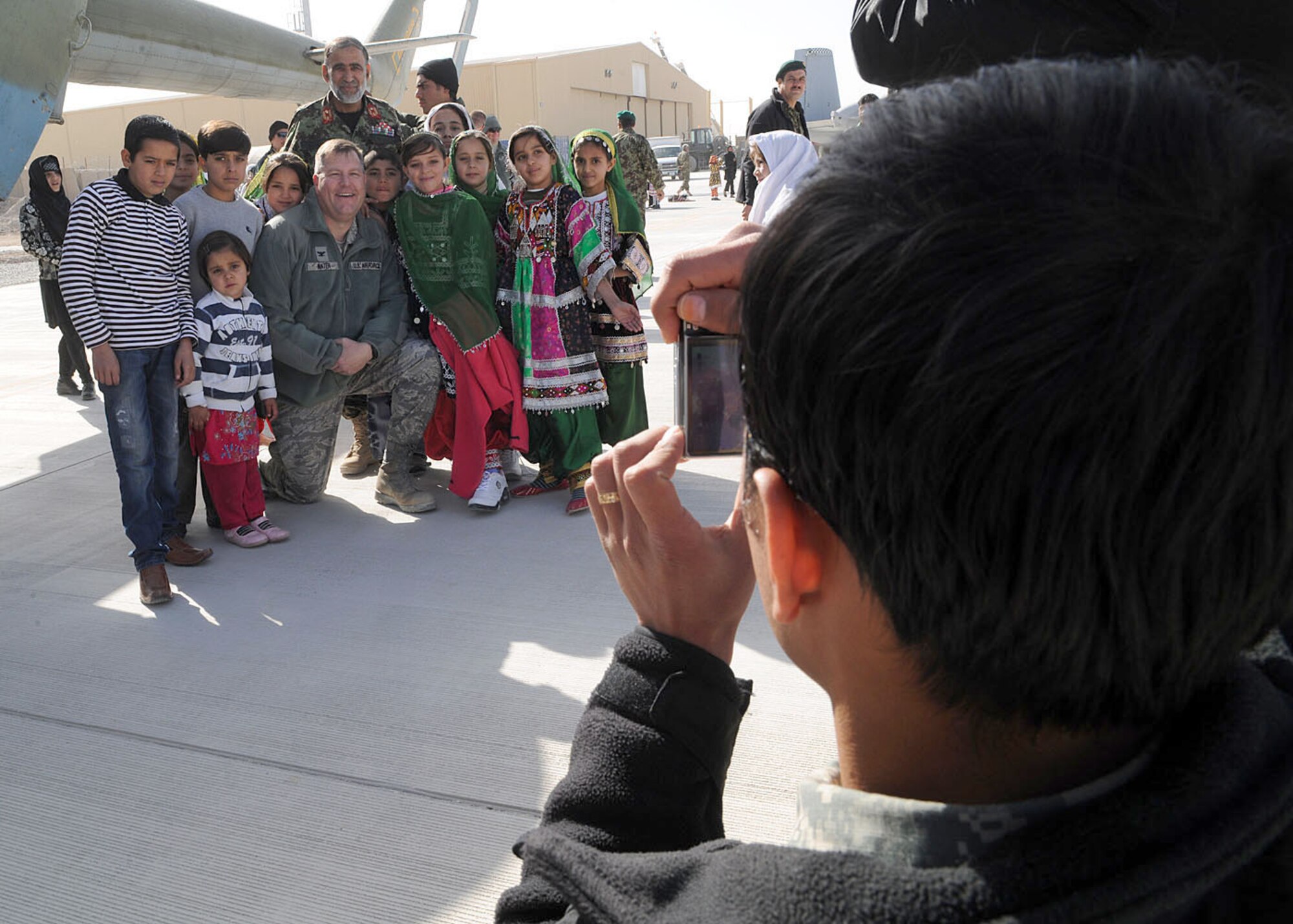 An Afghan citizen takes a photograph of school children posing with Col. Bernard Mater and Afghan air force Brig. Gen. Mohammed Yousuf during the Kandahar Air Wing open house Jan. 1, 2011, at Kandahar Airfield, Afghanistan. The open house showcased the newly constructed facilities and the inventory of the force. Coalition forces, tribal elders and school children attended the event to foster the continued partnership between the Afghan national security forces and International Assistance Security Force. Colonel Mater is the 738th Air Expeditionary Advisory Group commander, and General Yousuf is the Kandahar AW commander. (U.S. Air Force photo/Senior Airman Daryl Knee)
