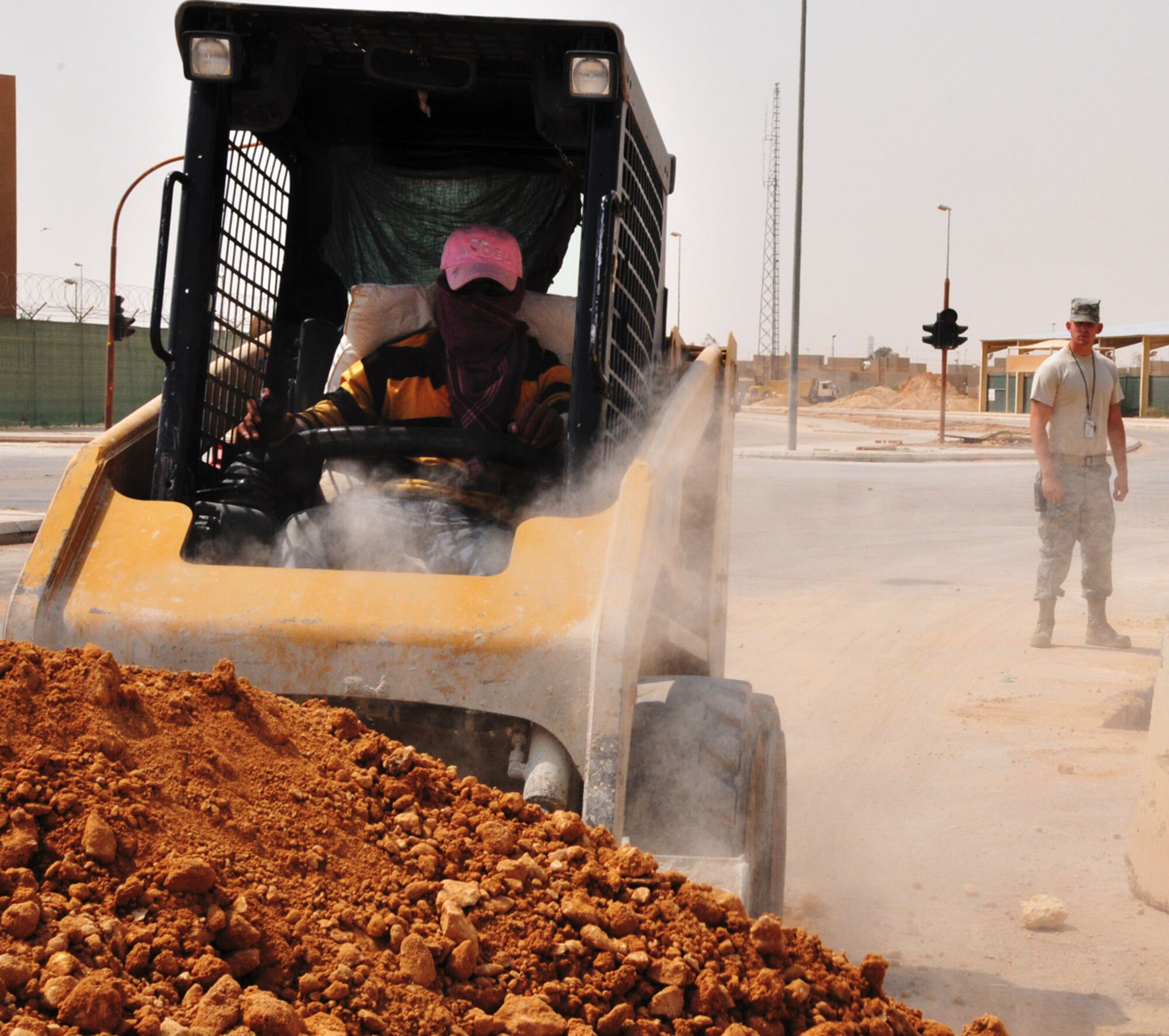 A local contract worker picks up soil to fill a trench after laying a new water line in the ground at a deployed location in Southwest Asia, June 28, as Senior Airman Patrick Meyers looks on. Meyers, a third country national escort deployed to the 64th Expeditionary Support Squadron's civil engineer escort flight, is one of nearly two dozen military members working with host nation contractors to complete a multi-year, $4.7 million upgrade to the base's water system. (U.S. Air Force photo/Master Sgt. Mike Hammond)